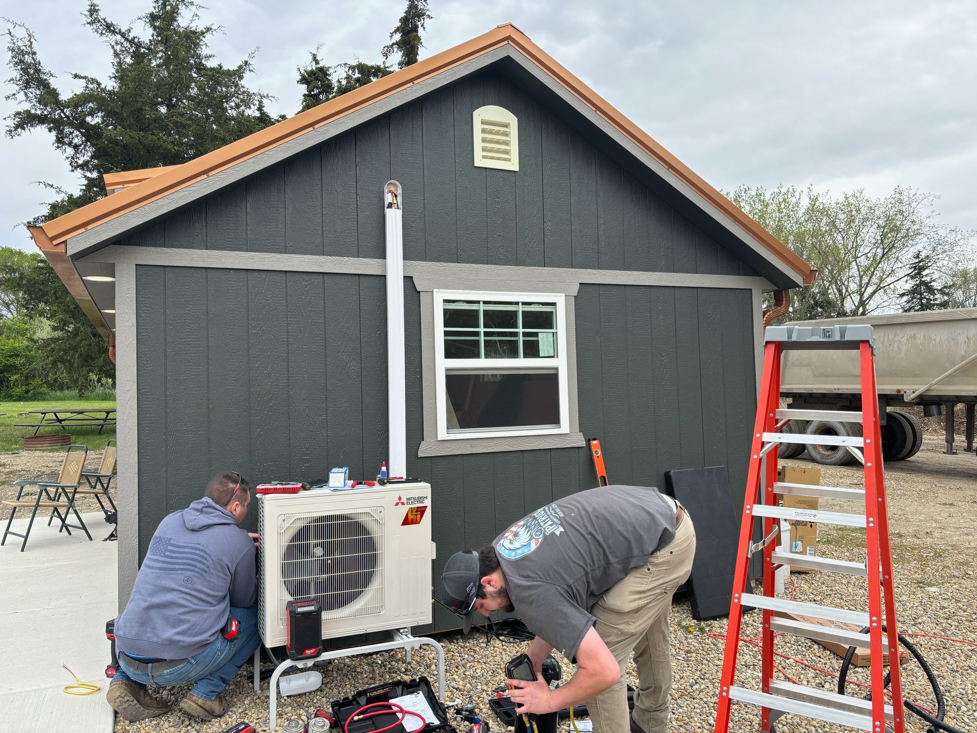 Two workers install a mini-split HVAC unit outside a dark-gray shed with a window and a nearby red ladder.