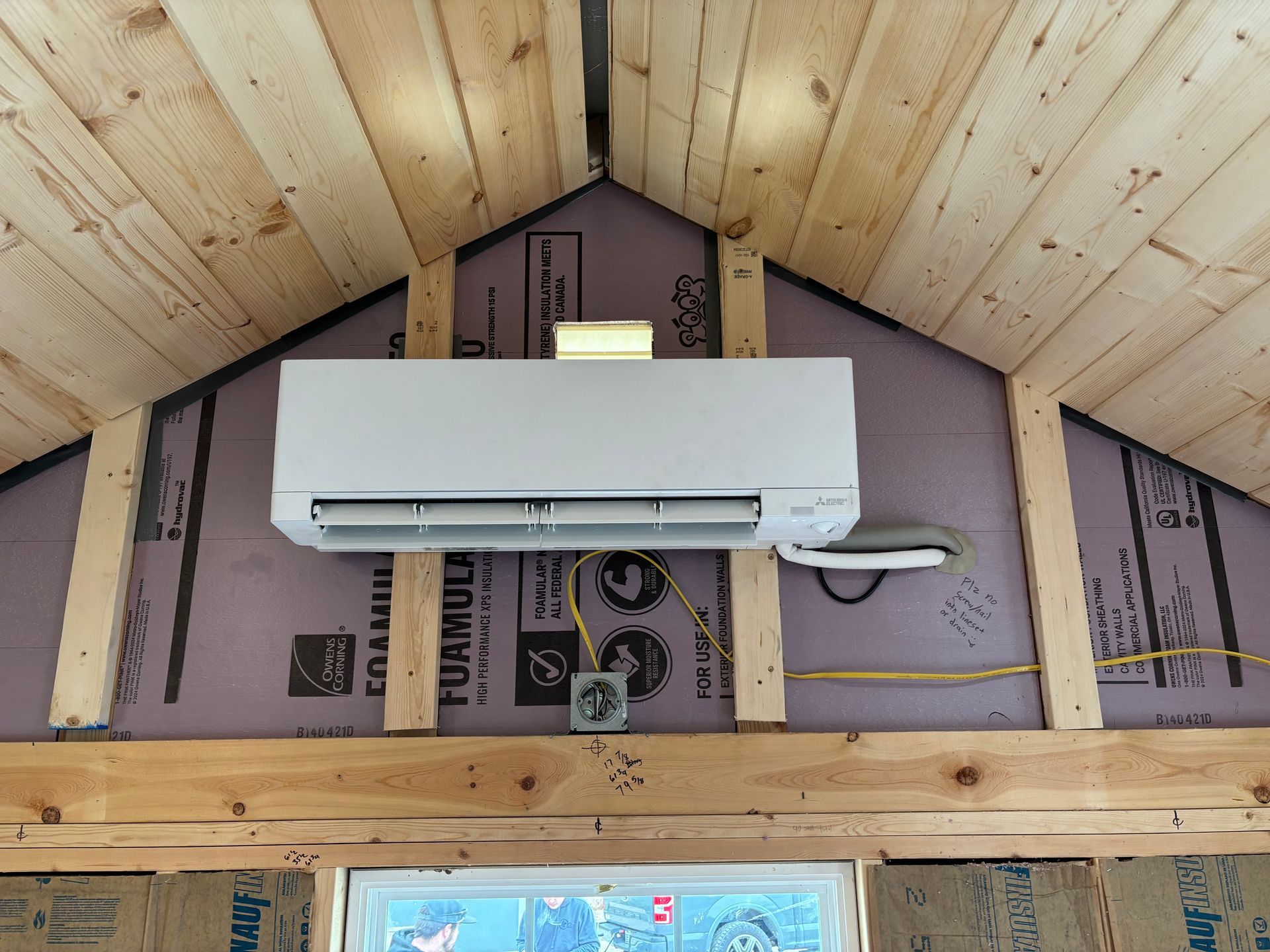 A white ductless mini-split air conditioner mounted on an unfinished wall with exposed wood studs and purple foam board.