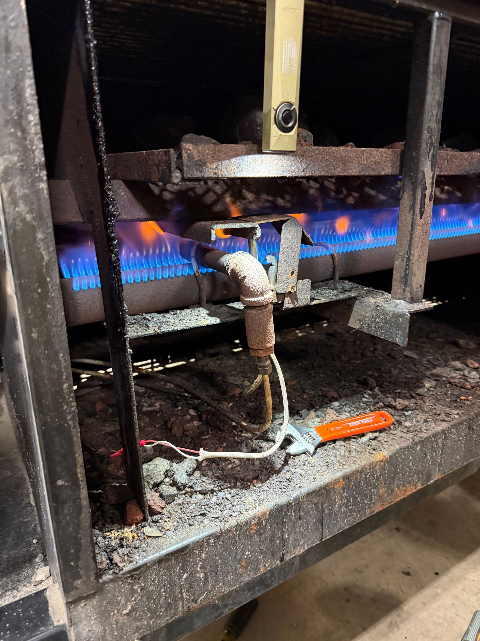 A lit gas burner unit inside an appliance, with a white wire leading to an orange-handled tool resting on the floor.
