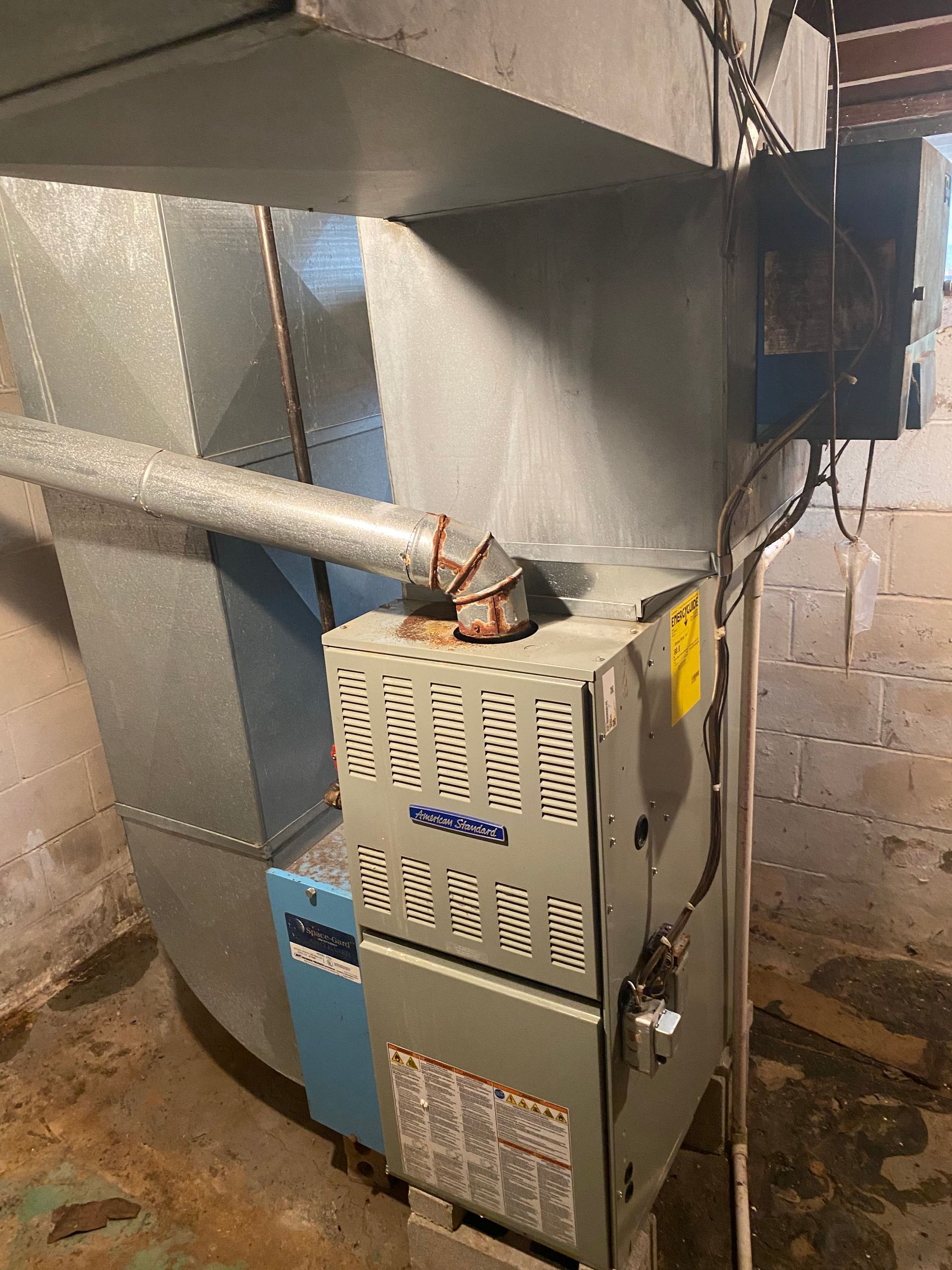 A residential HVAC furnace unit with metal ductwork and a connected vent pipe in a basement setting.