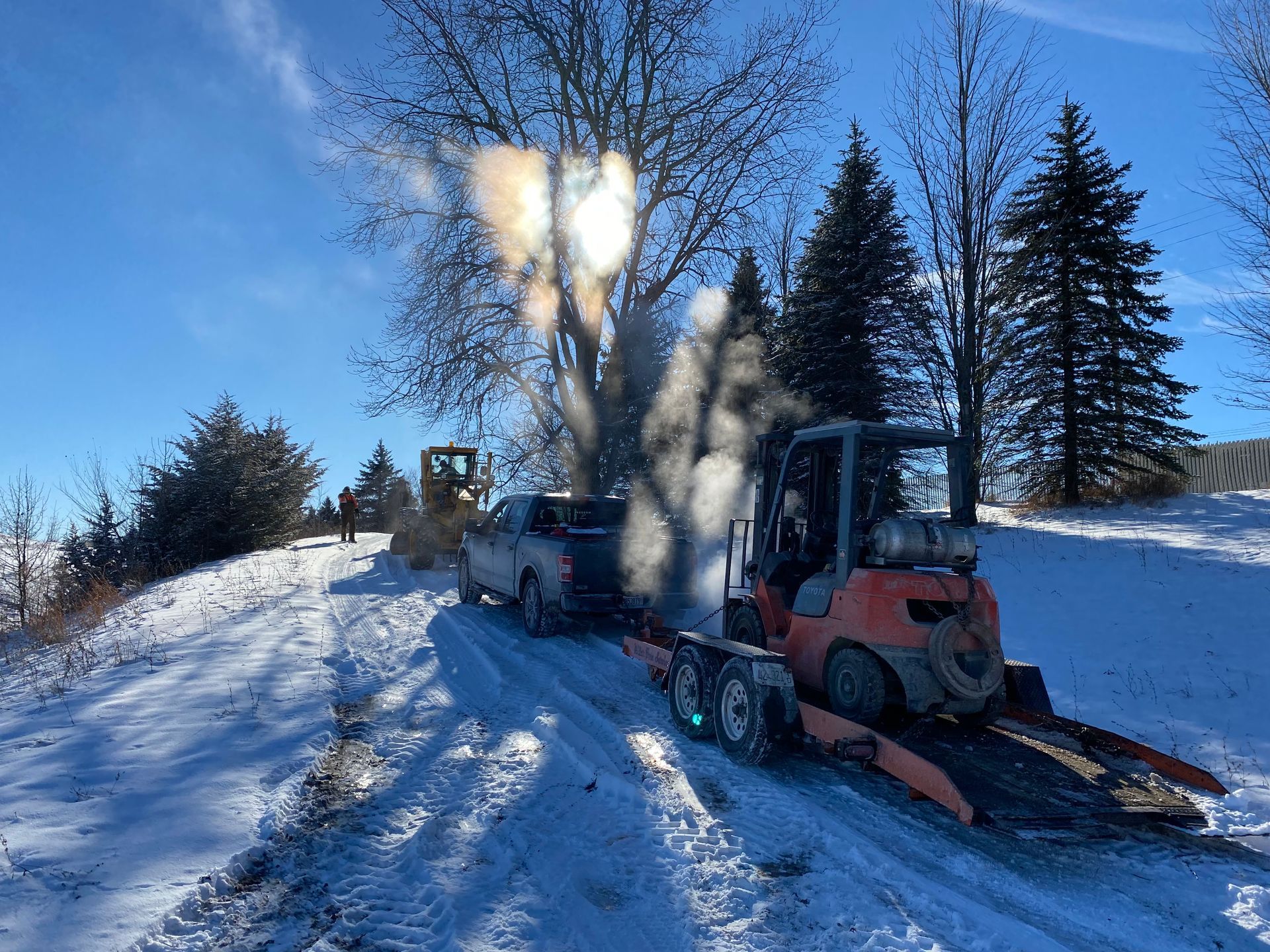 A pickup truck towing a flatbed trailer with an orange forklift is parked on a snowy, sunny landscape.