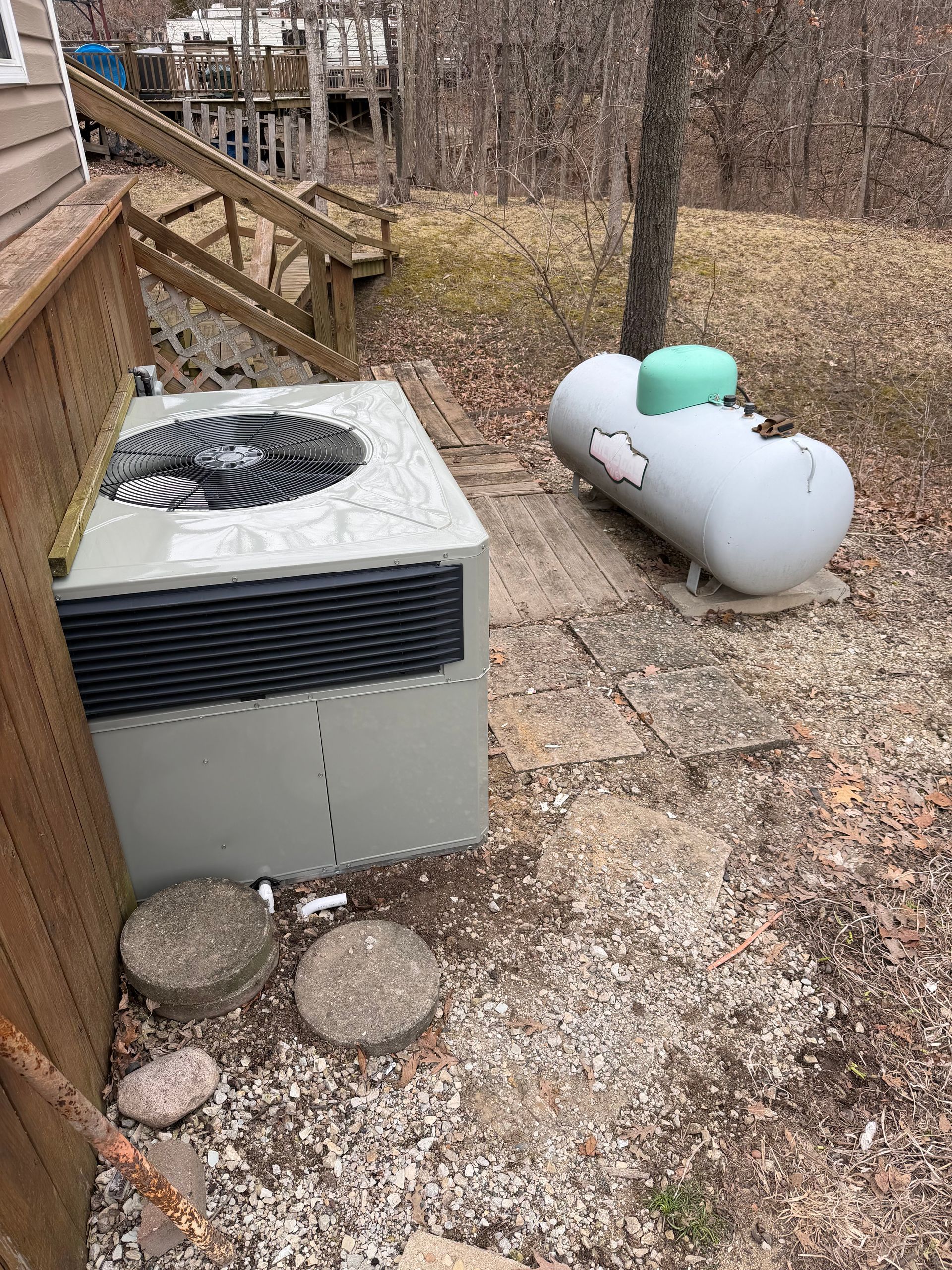 An outdoor HVAC unit next to a light gray propane tank on a gravel surface near a wooden deck and wooded area.