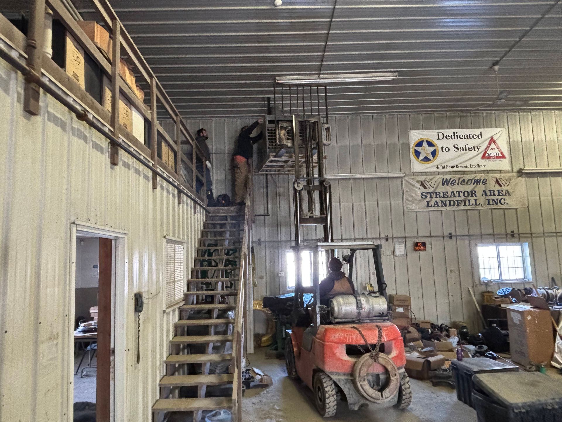 A forklift operator lifts a person near a mezzanine in a warehouse with corrugated metal walls.