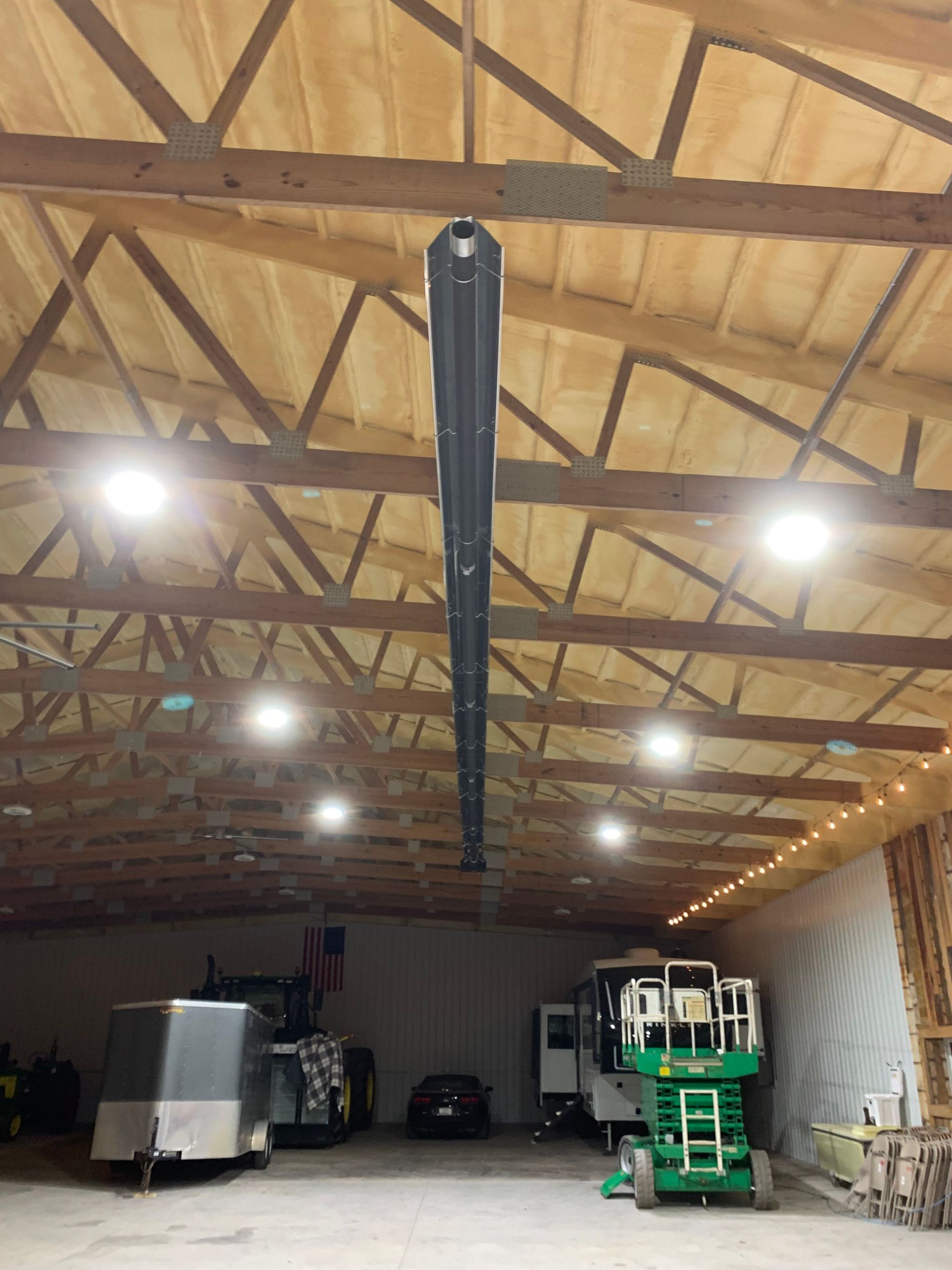 A large warehouse interior featuring a long overhead tube heater, bright round lights, a trailer, and a scissor lift.