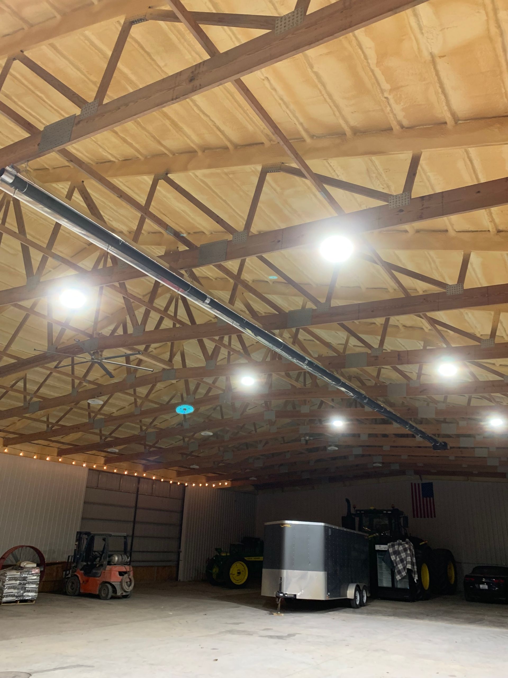 An interior view of a large, well-lit barn storing a forklift, a trailer, farm machinery, and an American flag.
