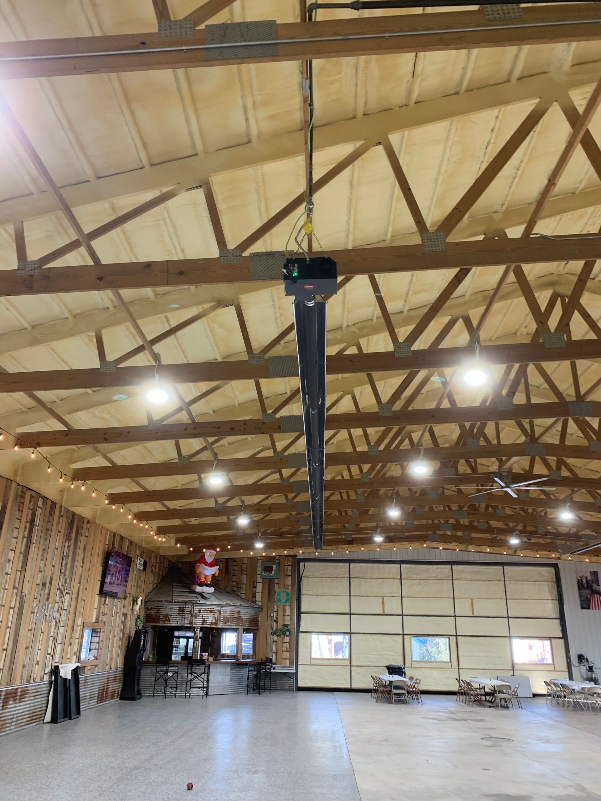 A spacious hall with exposed wooden trusses, recessed ceiling lights, a hanging tube heater, and a bar area in the back.