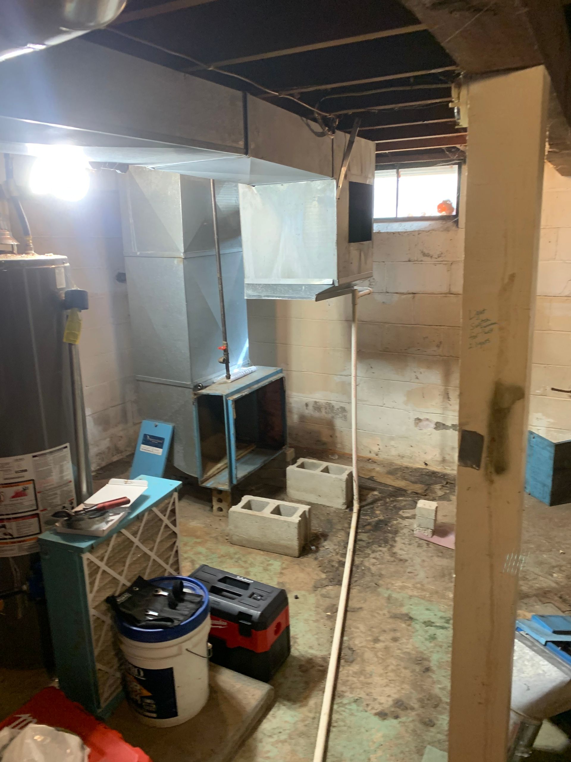 A basement renovation in progress showing HVAC ductwork, a water heater, a bucket, tools, and cinder blocks on the floor.