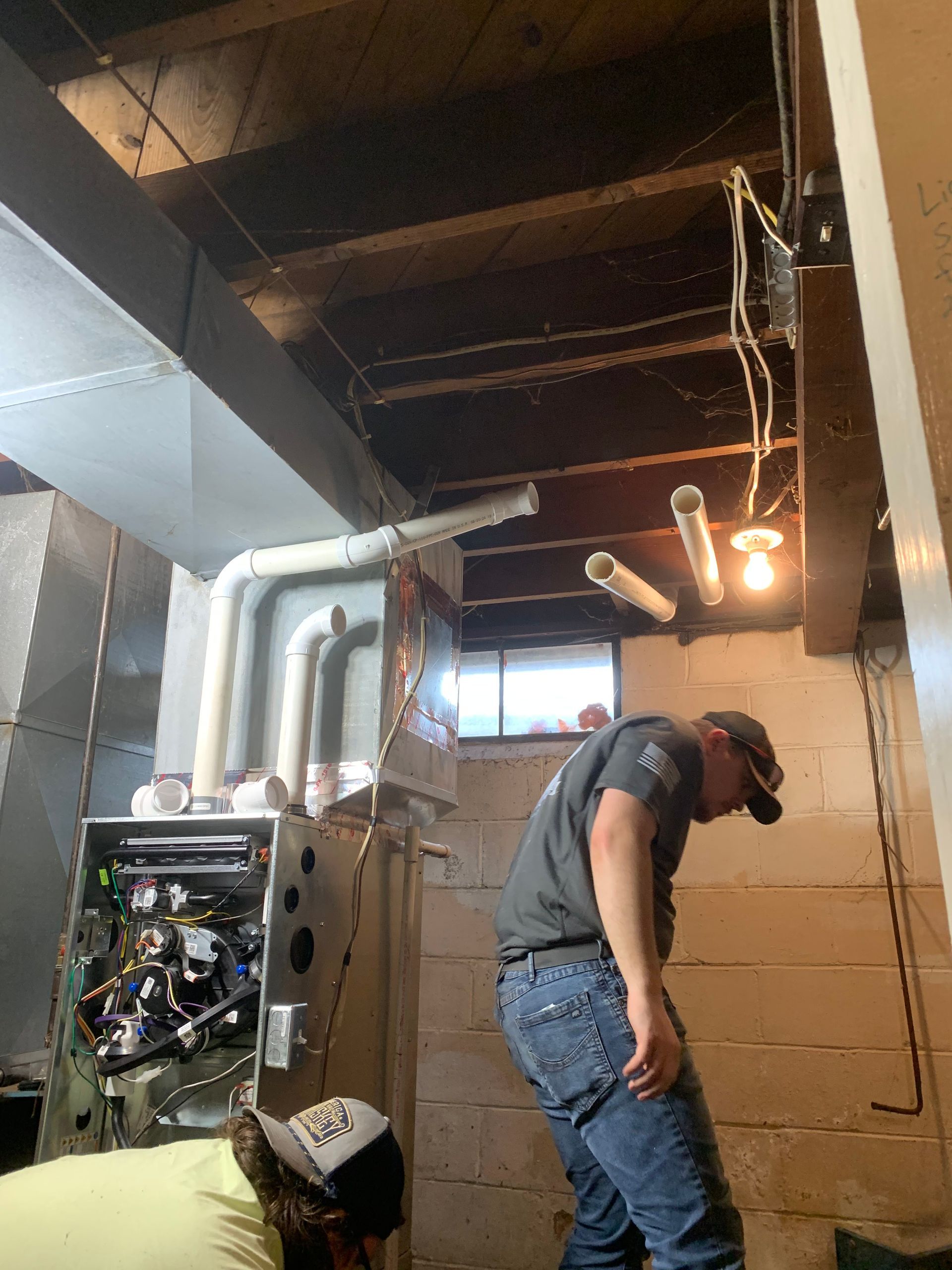 Two workers in a basement installing or servicing a residential HVAC furnace unit with exposed white PVC venting pipes.