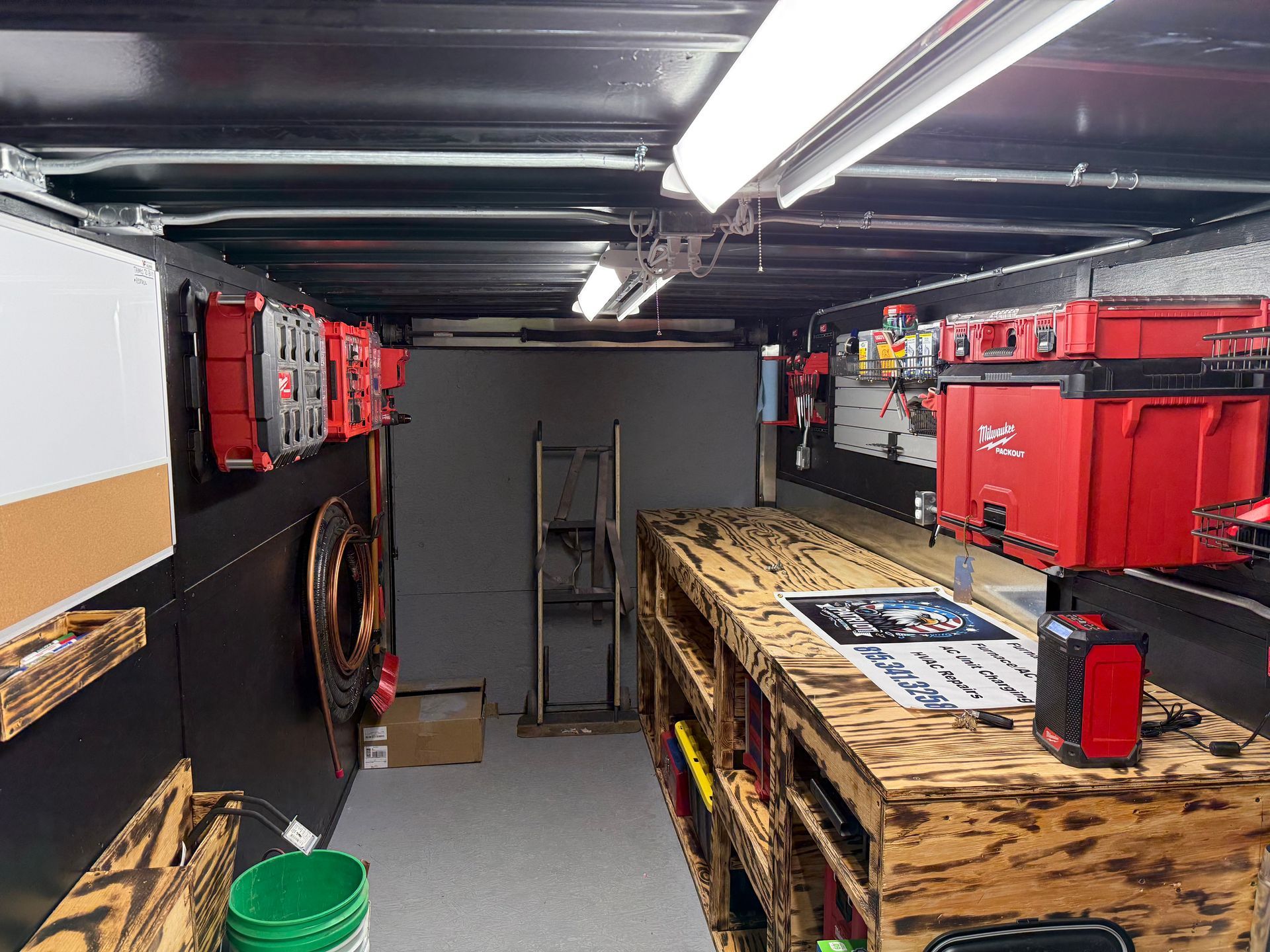Interior view of a work trailer featuring wooden storage shelving on the right, wall-mounted red tool cases, and a ramp.