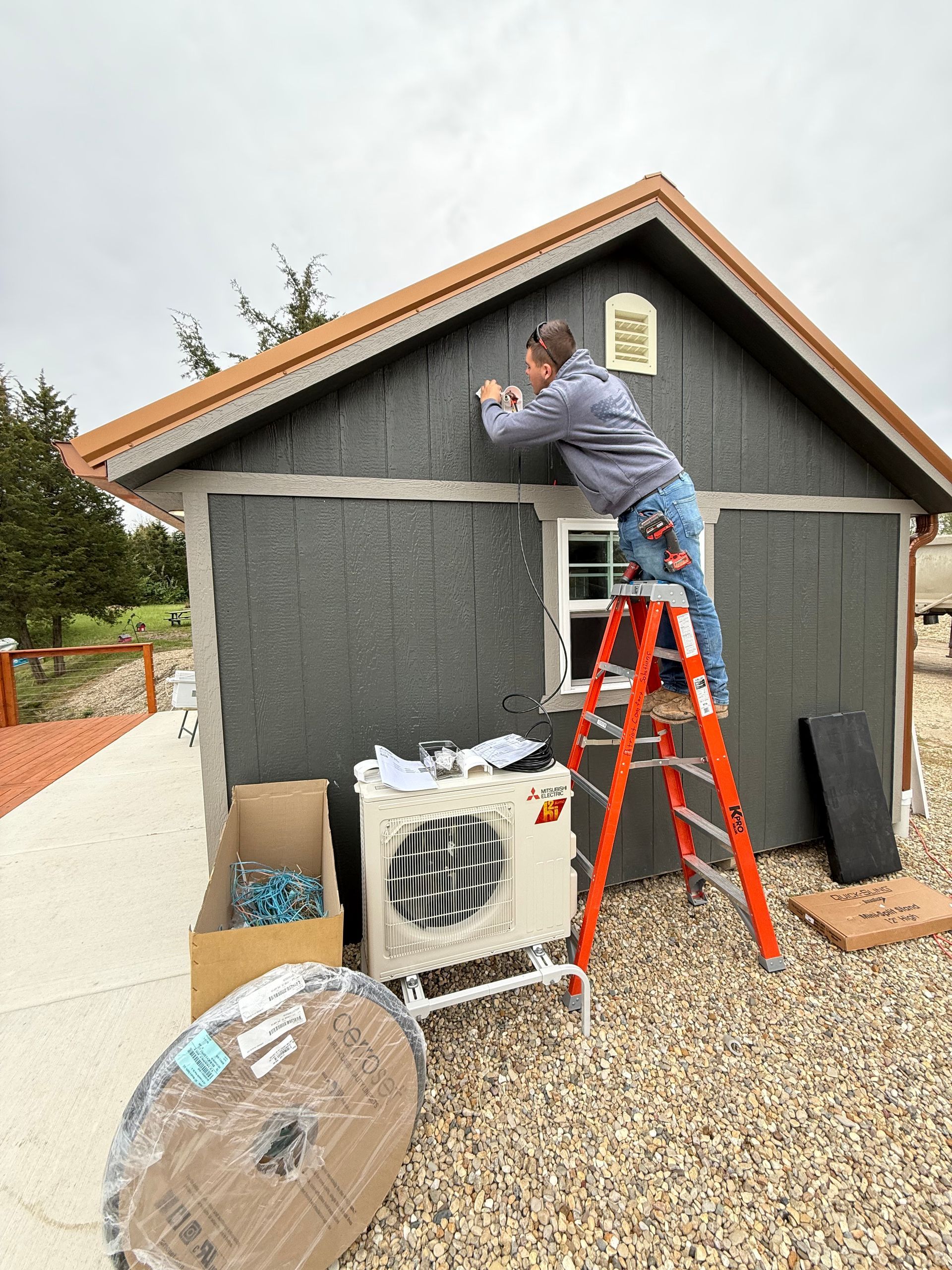 A person standing on an orange ladder installs hardware on the dark gray siding of a small building near an AC unit.