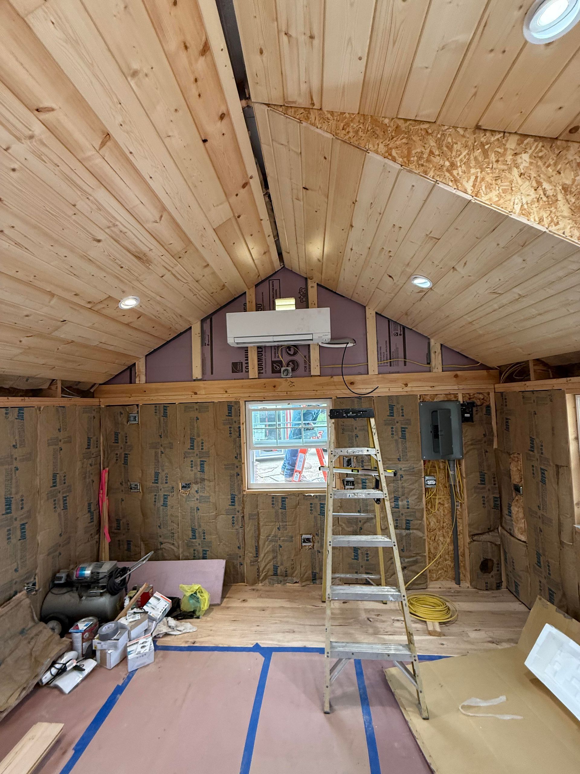 An interior view of a room under construction with exposed wooden rafters, insulation, a mini-split, and a step ladder.