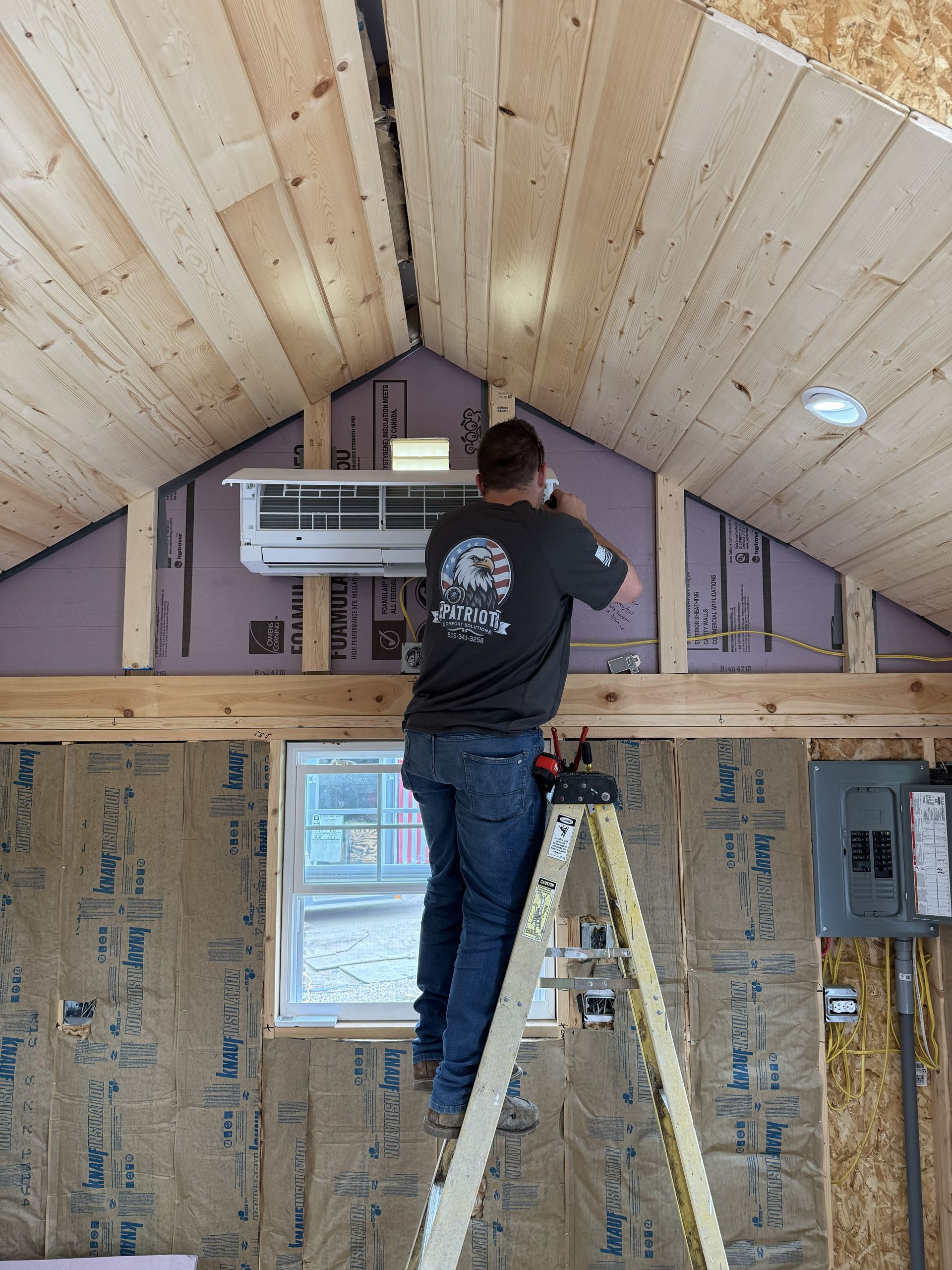 A person stands on a ladder, installing a wall-mounted ductless air conditioning unit in a room with unfinished walls.