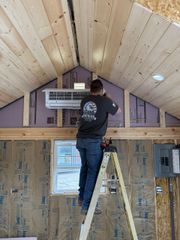 A person stands on a ladder, installing a wall-mounted ductless air conditioning unit in a room with unfinished walls.