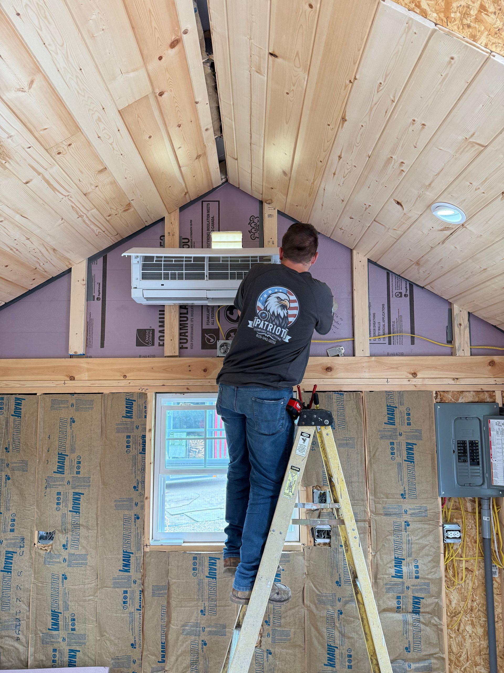 A person stands on a ladder, installing a white mini-split air conditioner on a wall framed with wooden boards and insulation.