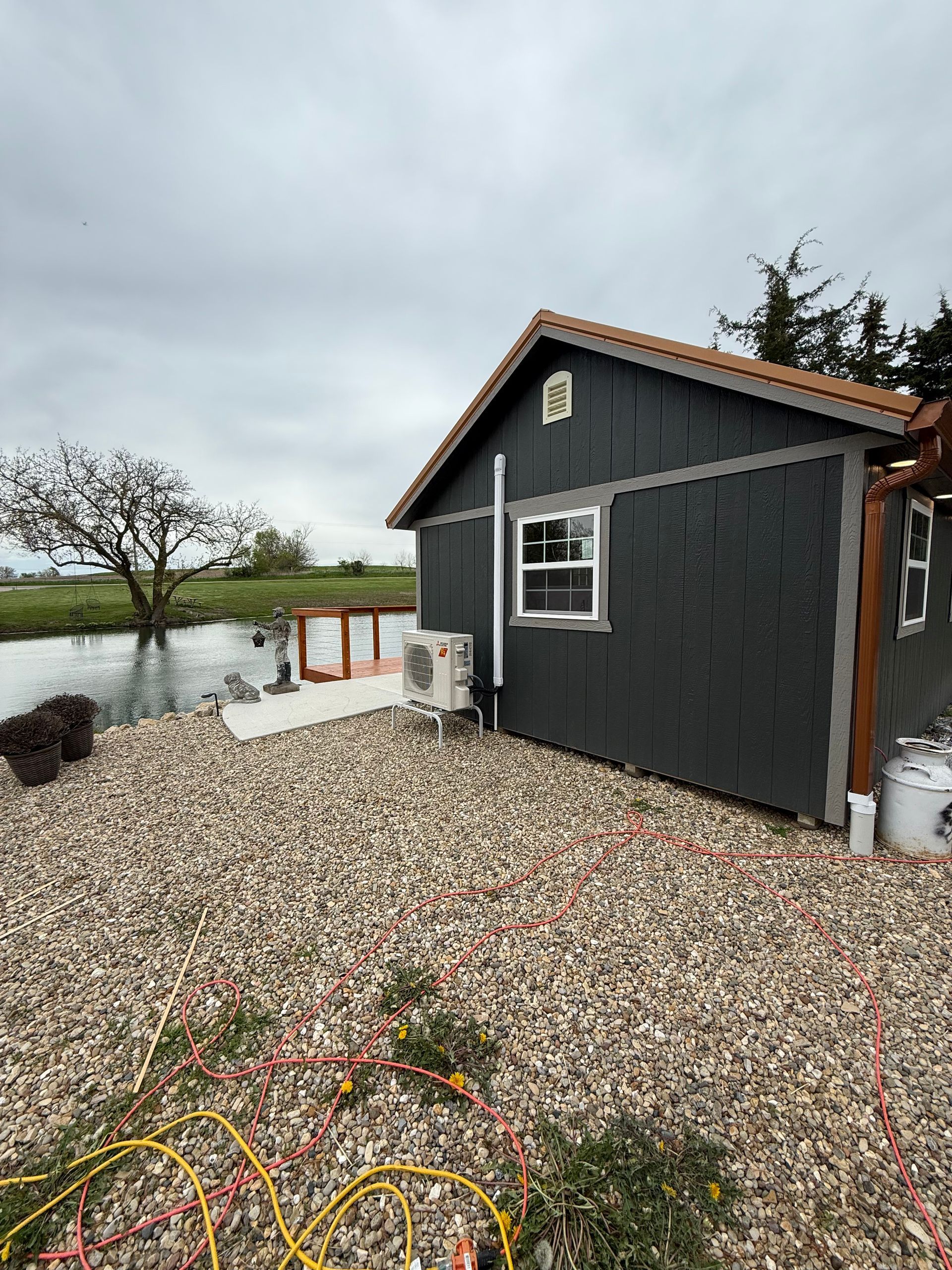 A gray, shingled shed with a window and HVAC unit sits on a gravel lot beside a stream under a cloudy sky.