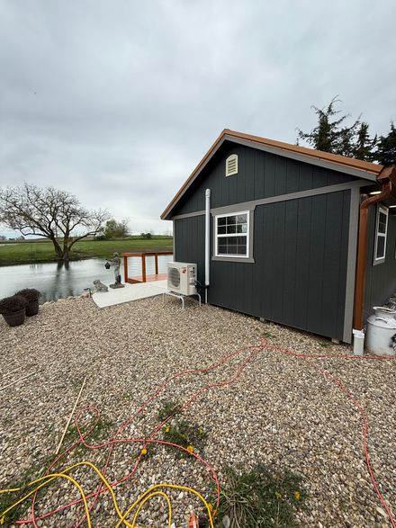 Dark gray shed with an outdoor HVAC unit and white-trimmed window, located on a gravel lot near a pond and grassy field.