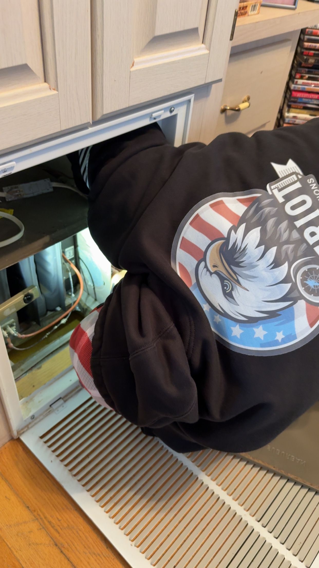 A person wearing a black eagle-print hoodie reaching into an open HVAC return vent near a floor.