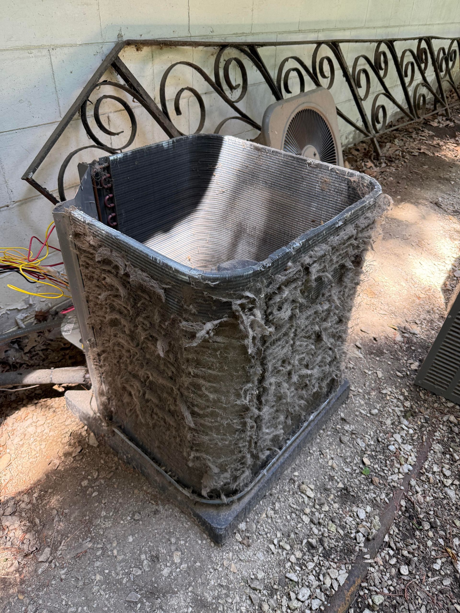 A dirty, square HVAC condenser coil covered in thick debris, sitting outdoors on a concrete base.