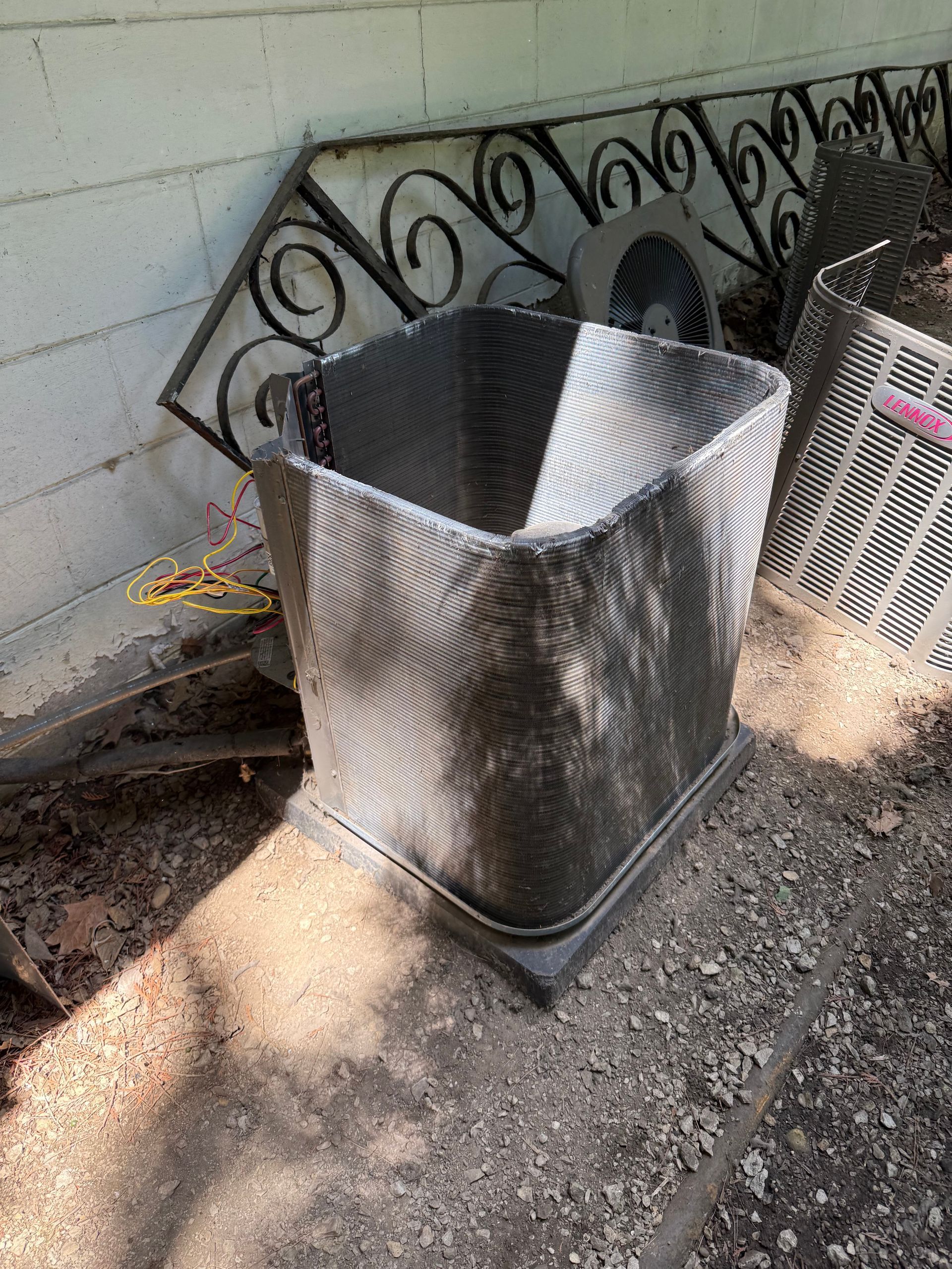 A metal air conditioning condenser coil sits outside on gravel, positioned in front of a white cinder block wall.