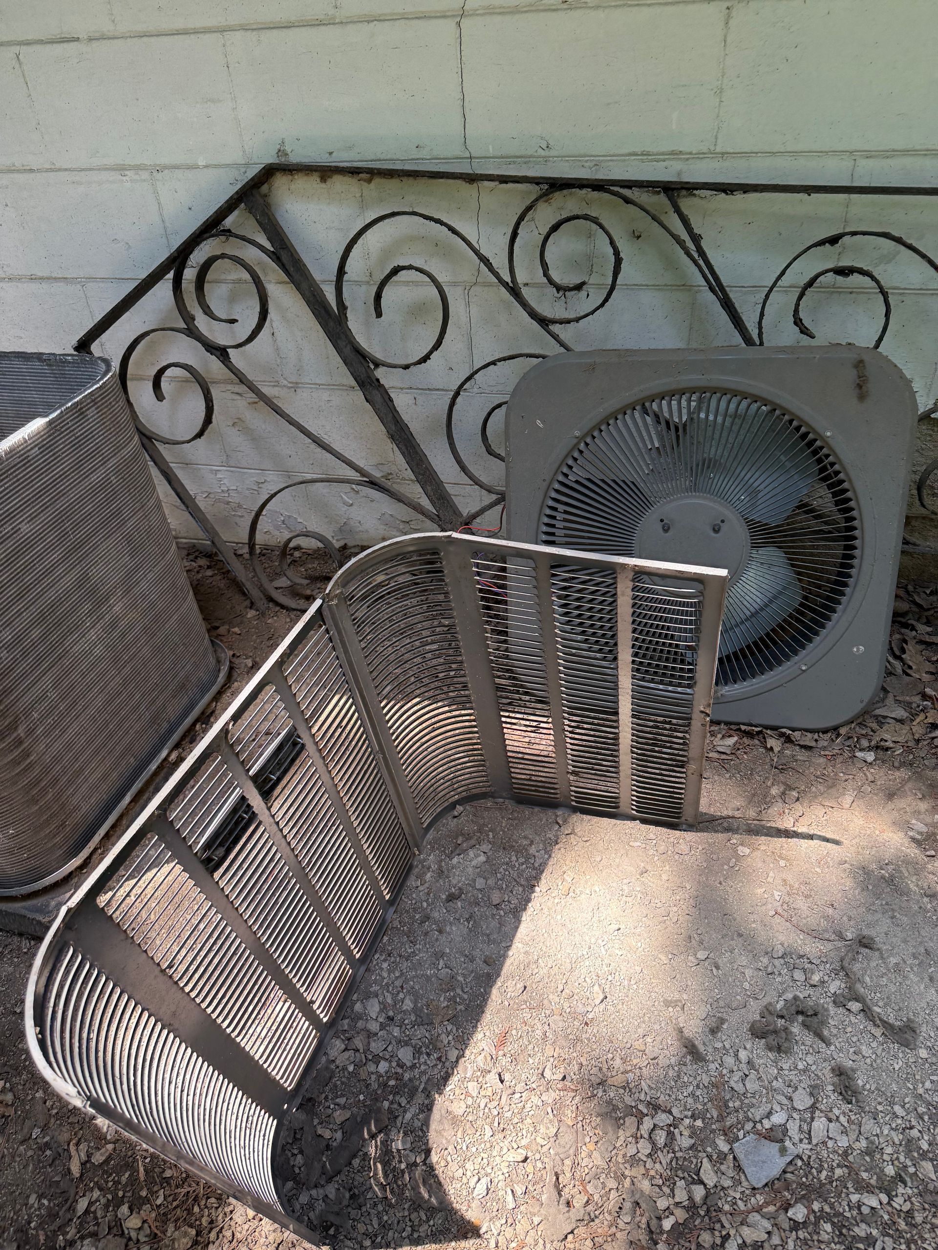 Discarded metal HVAC fan cover and decorative wrought iron railing resting against a painted cinder block wall.