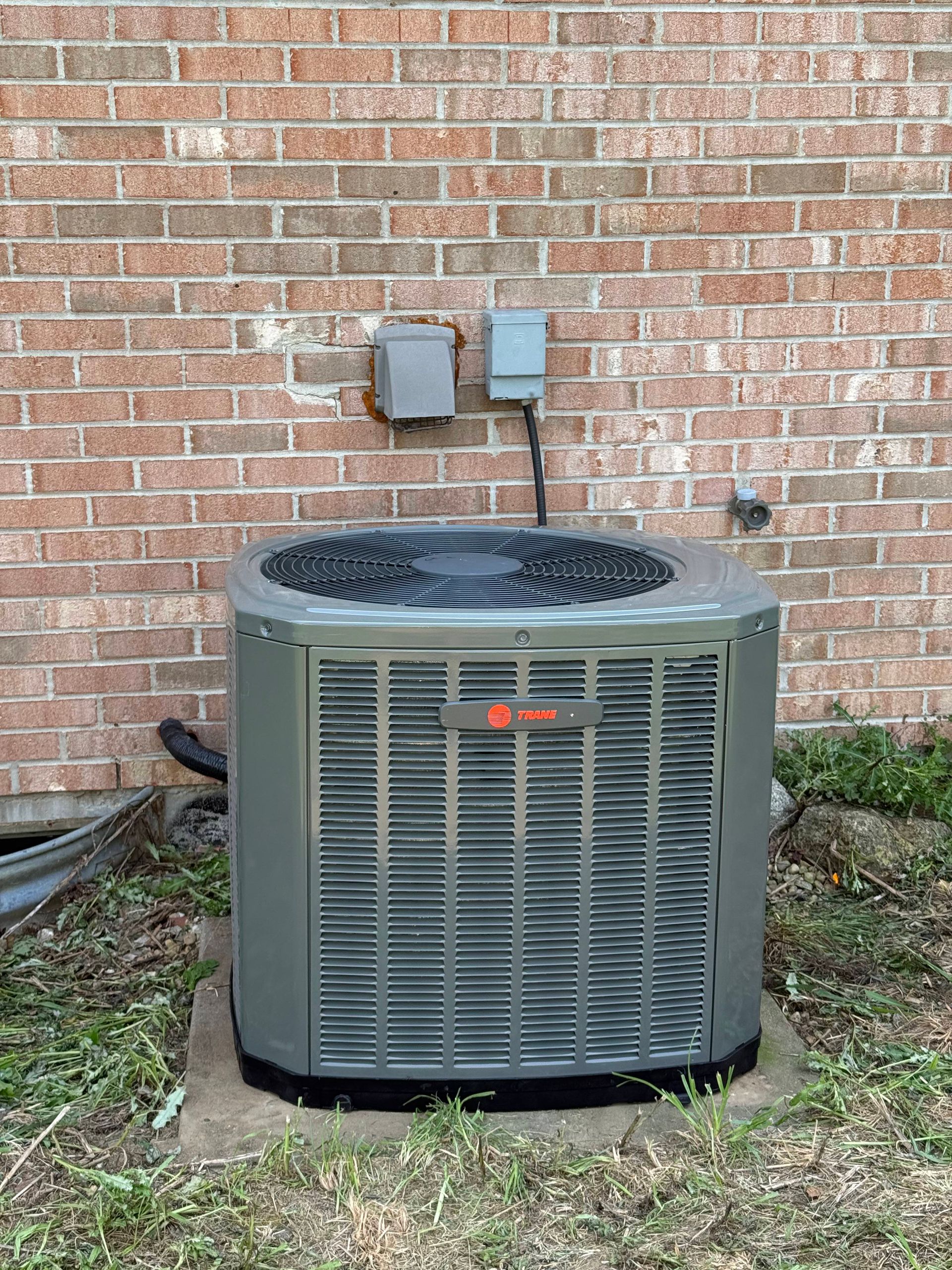 A gray Trane air conditioning unit sits on a concrete pad against a brick wall next to two electrical disconnect boxes.