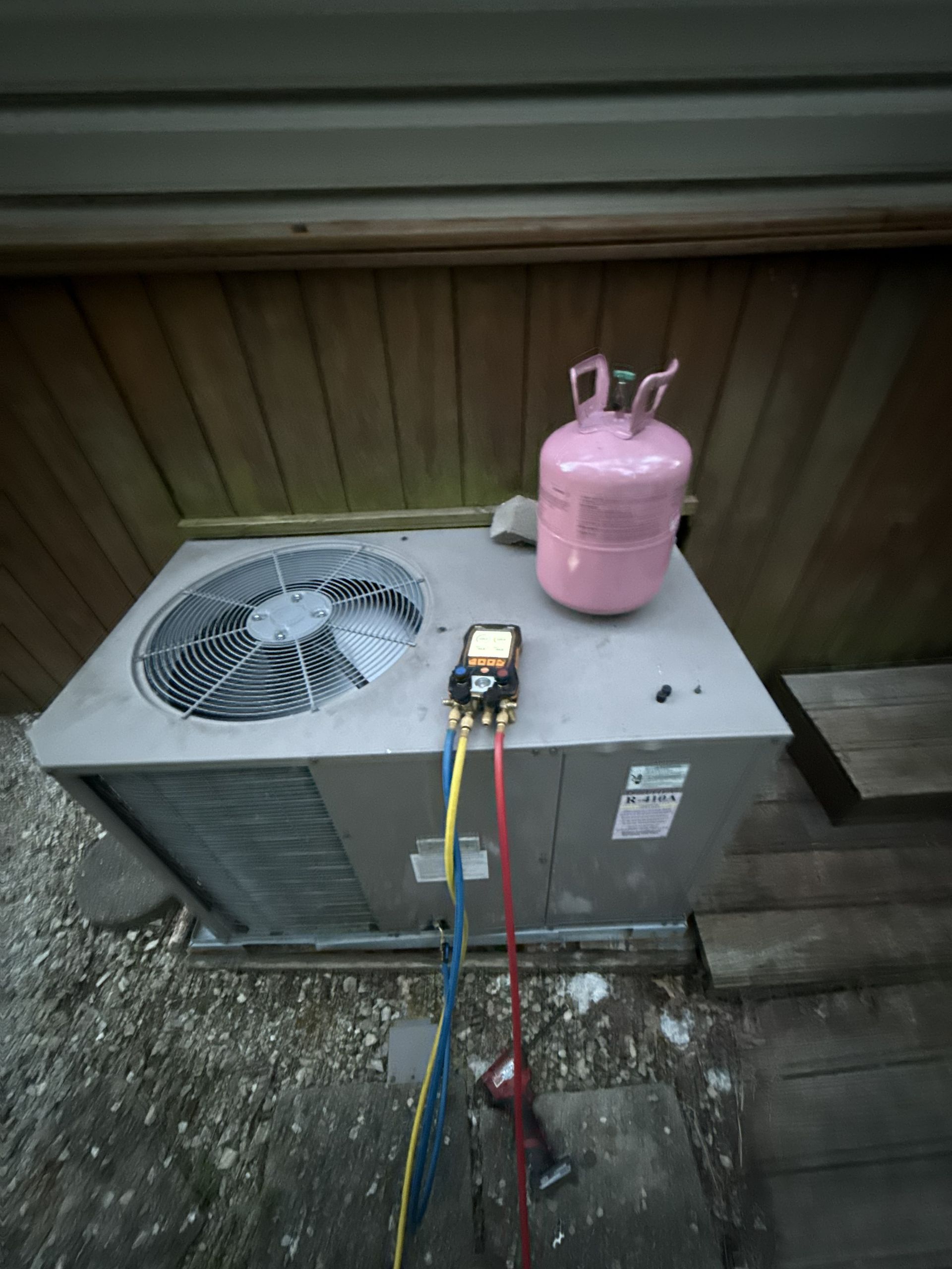 An outdoor air conditioning unit being serviced with a manifold gauge set and a pink refrigerant tank.