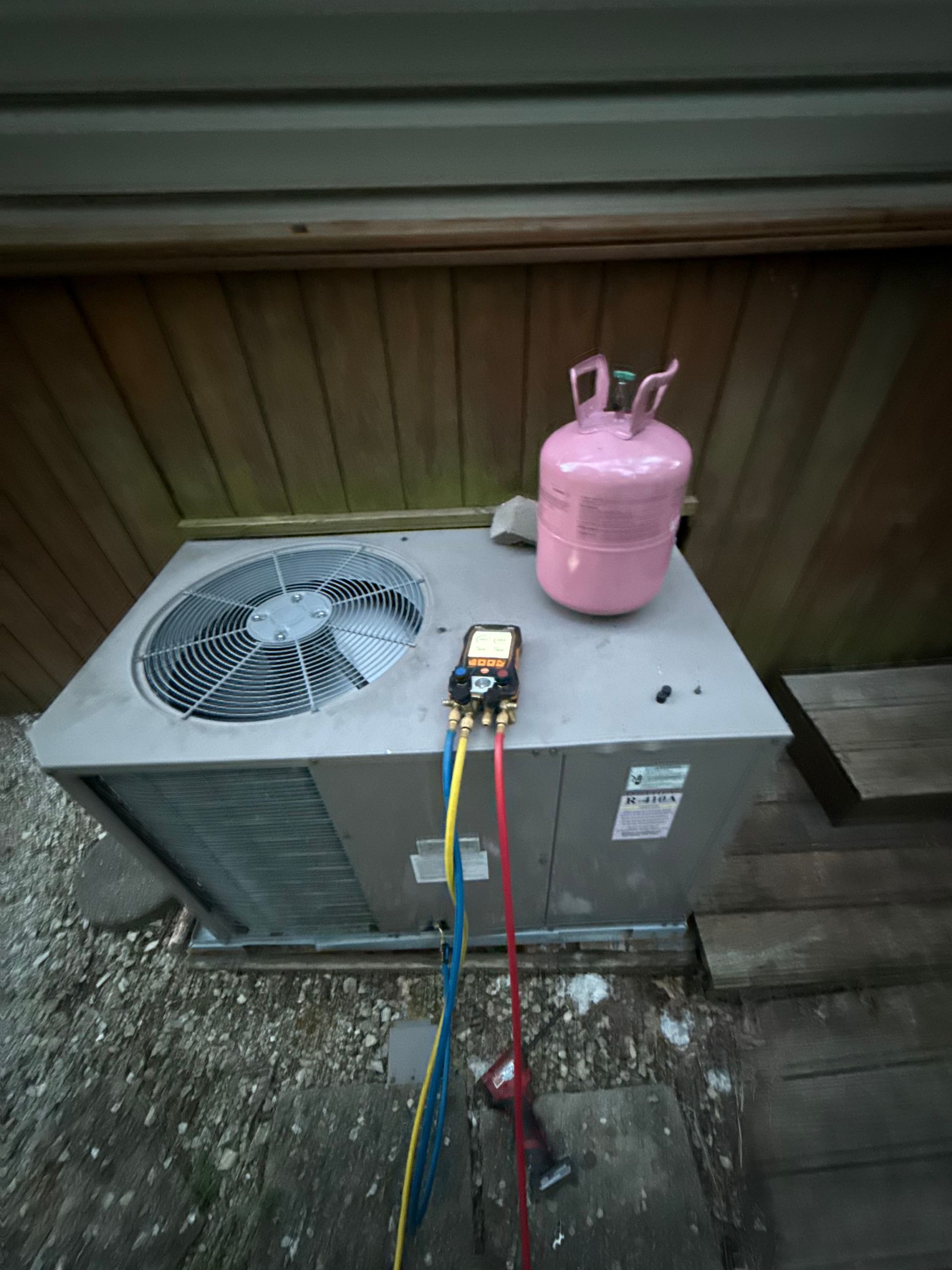An outdoor HVAC unit being serviced with a manifold gauge set connected to a pink refrigerant tank.