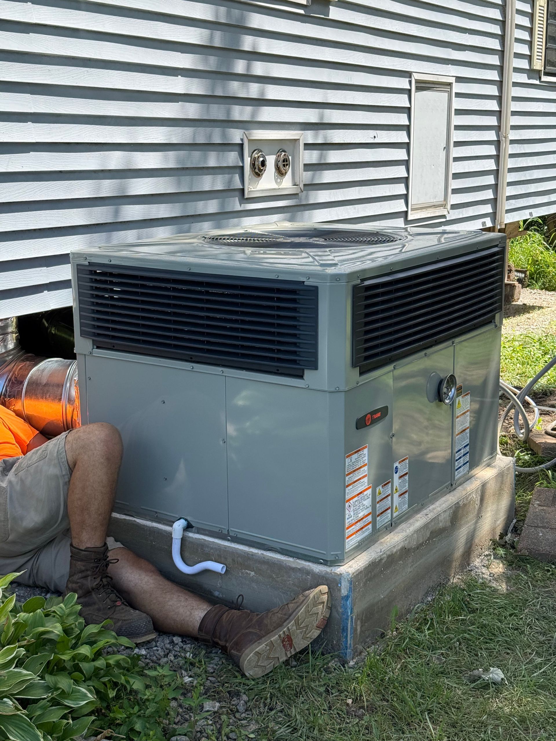 A technician in work boots and shorts kneeling on the ground while servicing an outdoor HVAC unit next to a house.