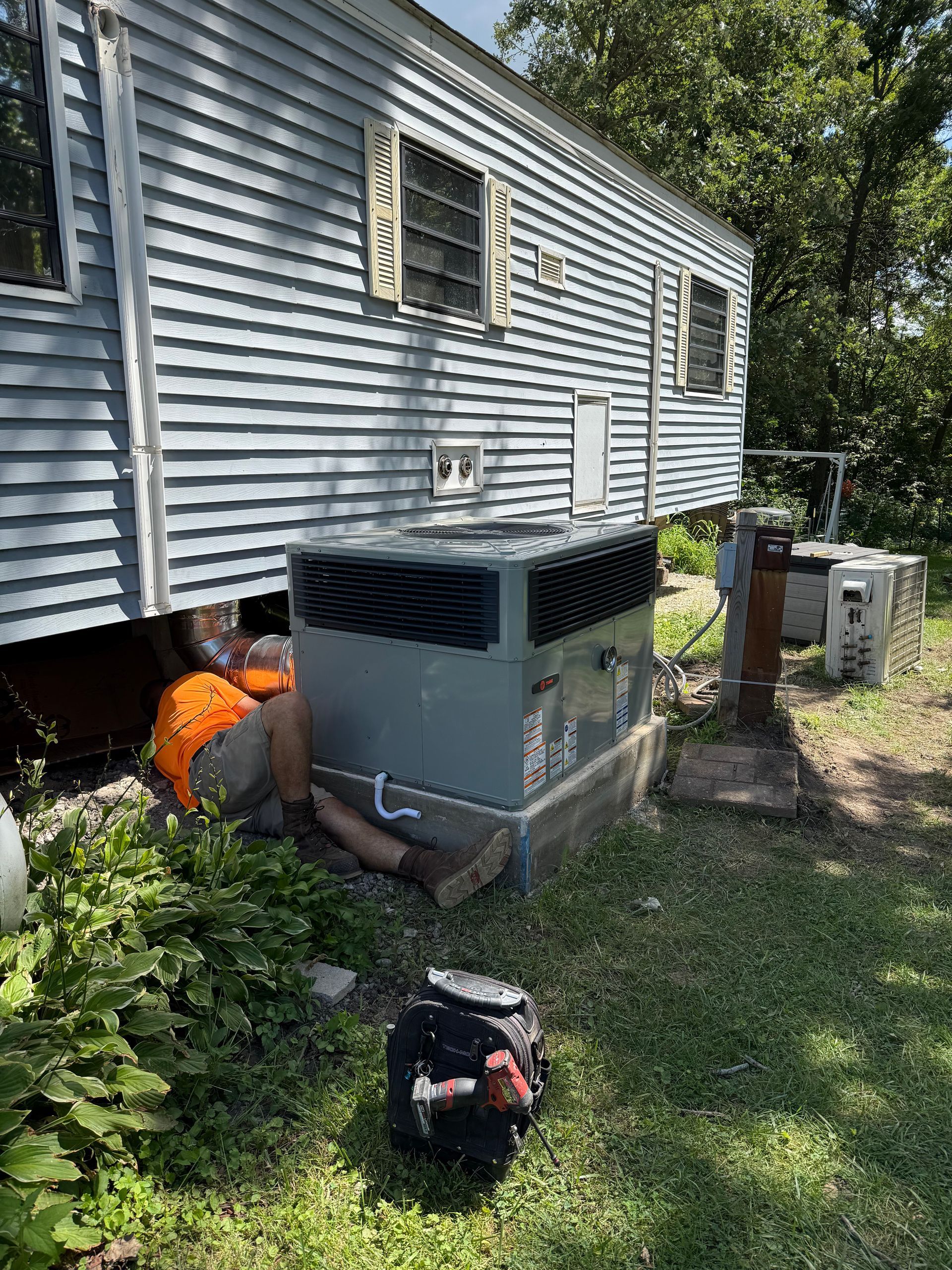 A technician in an orange shirt works on an air conditioning unit located next to a light-blue mobile home.
