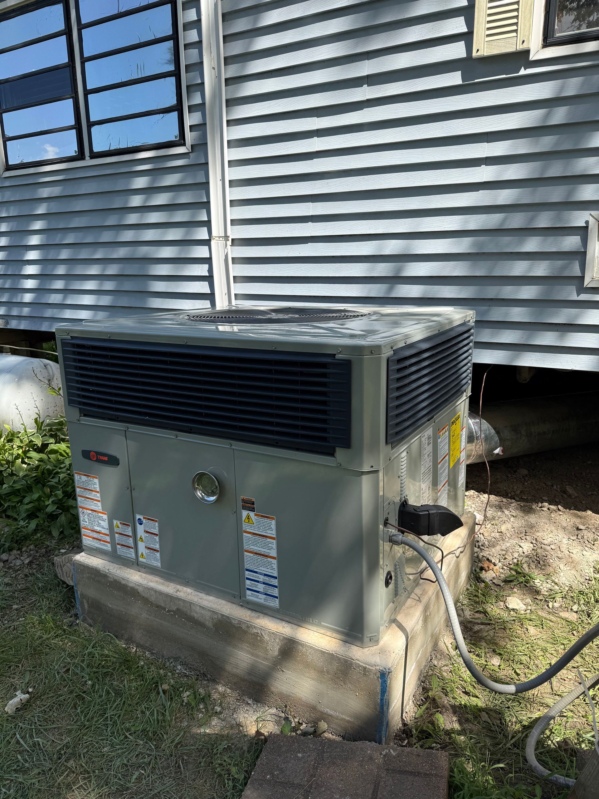 A gray HVAC packaged unit sits on a concrete pad outside a house with gray vinyl siding.