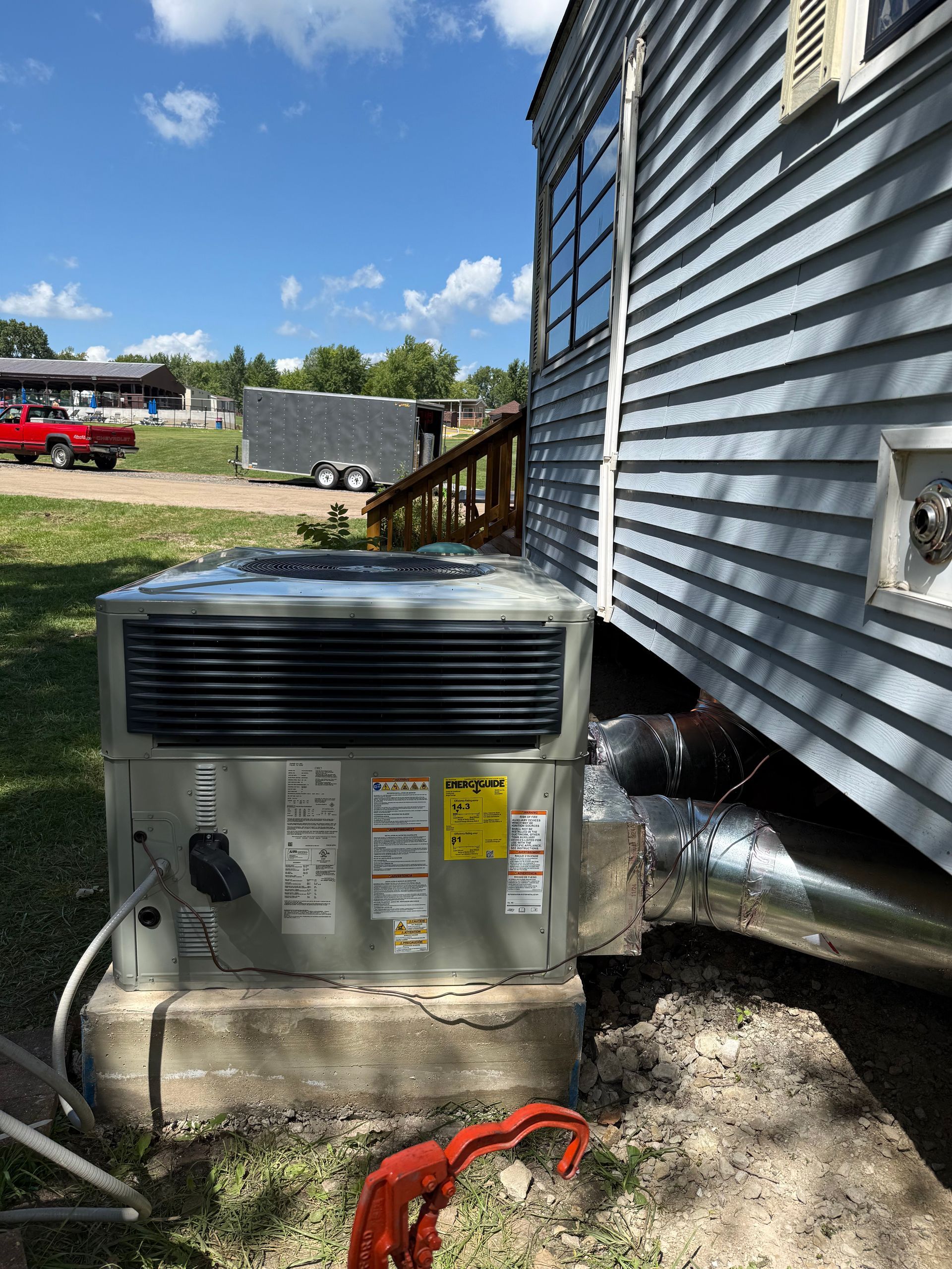 A grey HVAC unit sits on a concrete pad next to a light-sided mobile home, with a duct connection to the home's side.