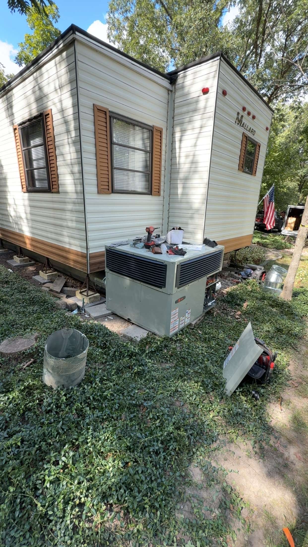 A light-colored mobile home exterior with a central air conditioning unit on the grass in front of a wall with shutters.