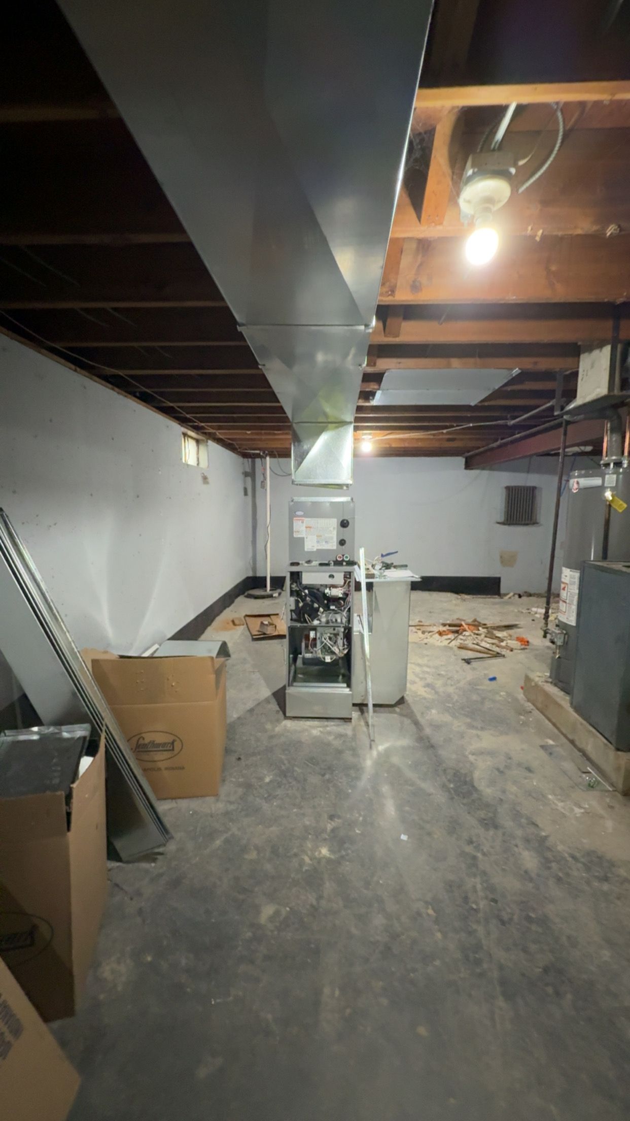 A basement interior featuring an HVAC furnace unit, exposed wooden ceiling joists, and a central metal duct run.