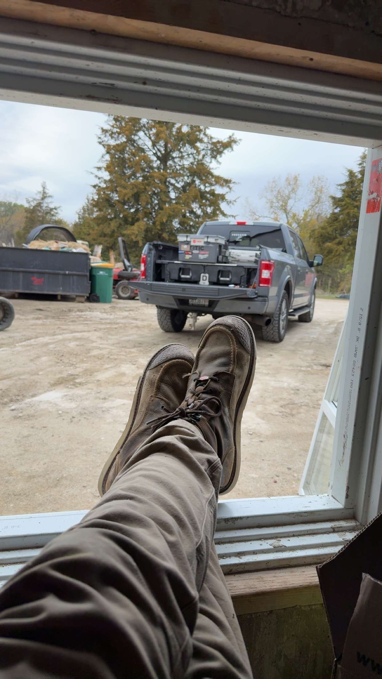 A person's legs, wearing boots and pants, rest on a window ledge looking out at a parked pickup truck on a dirt lot.