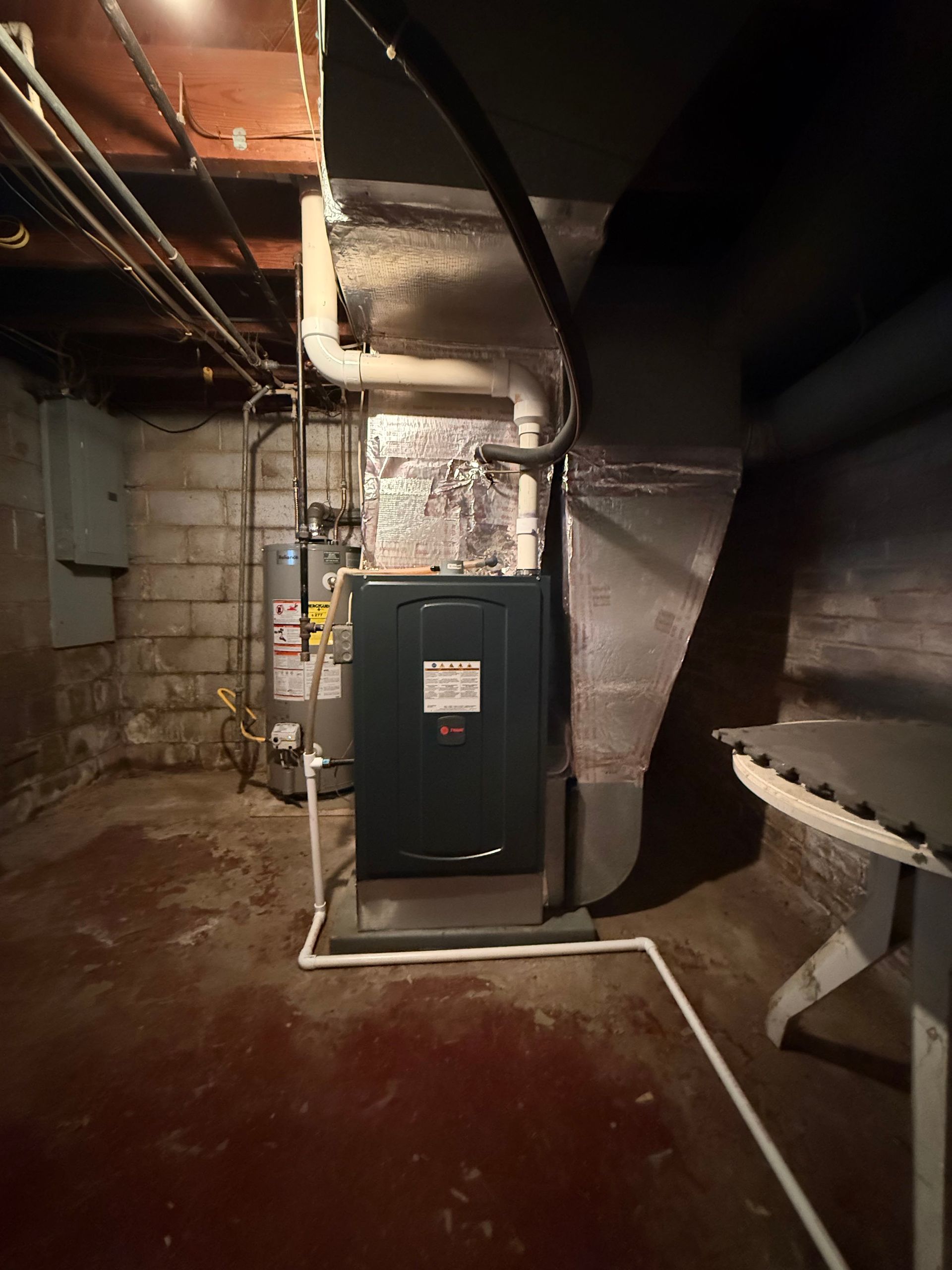 A furnace and water heater stand in a dim, unfinished basement with concrete block walls and exposed ceiling joists.