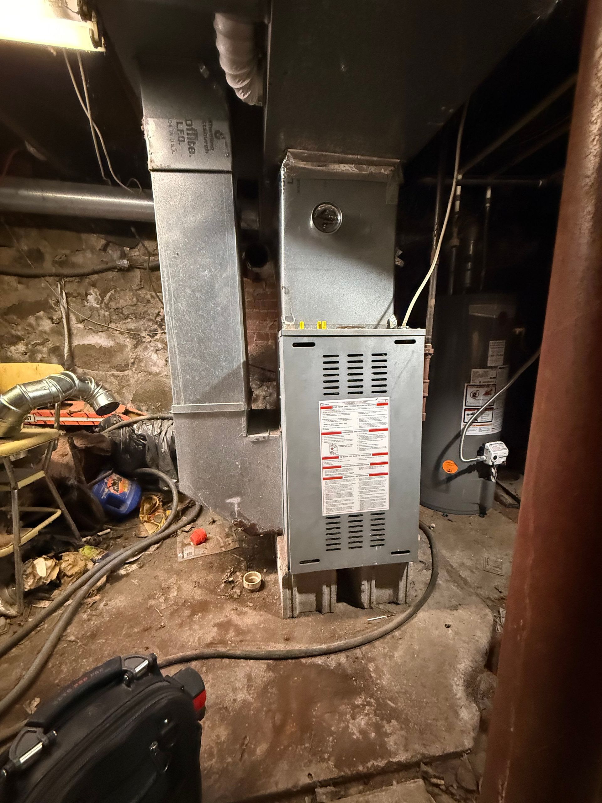 A furnace and ductwork system with a water heater in the background inside a basement with concrete walls and floor.