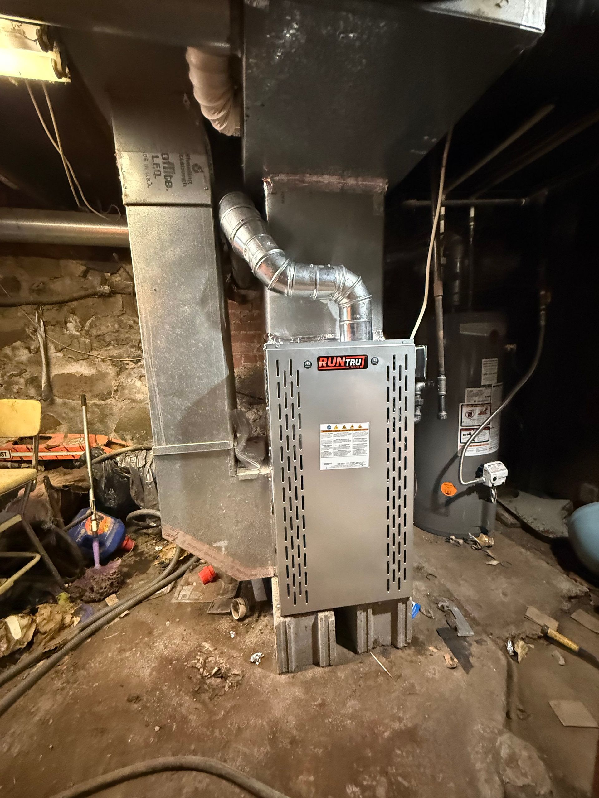 A silver metal humidifier unit attached to a vertical furnace duct in a dark basement near a water heater.