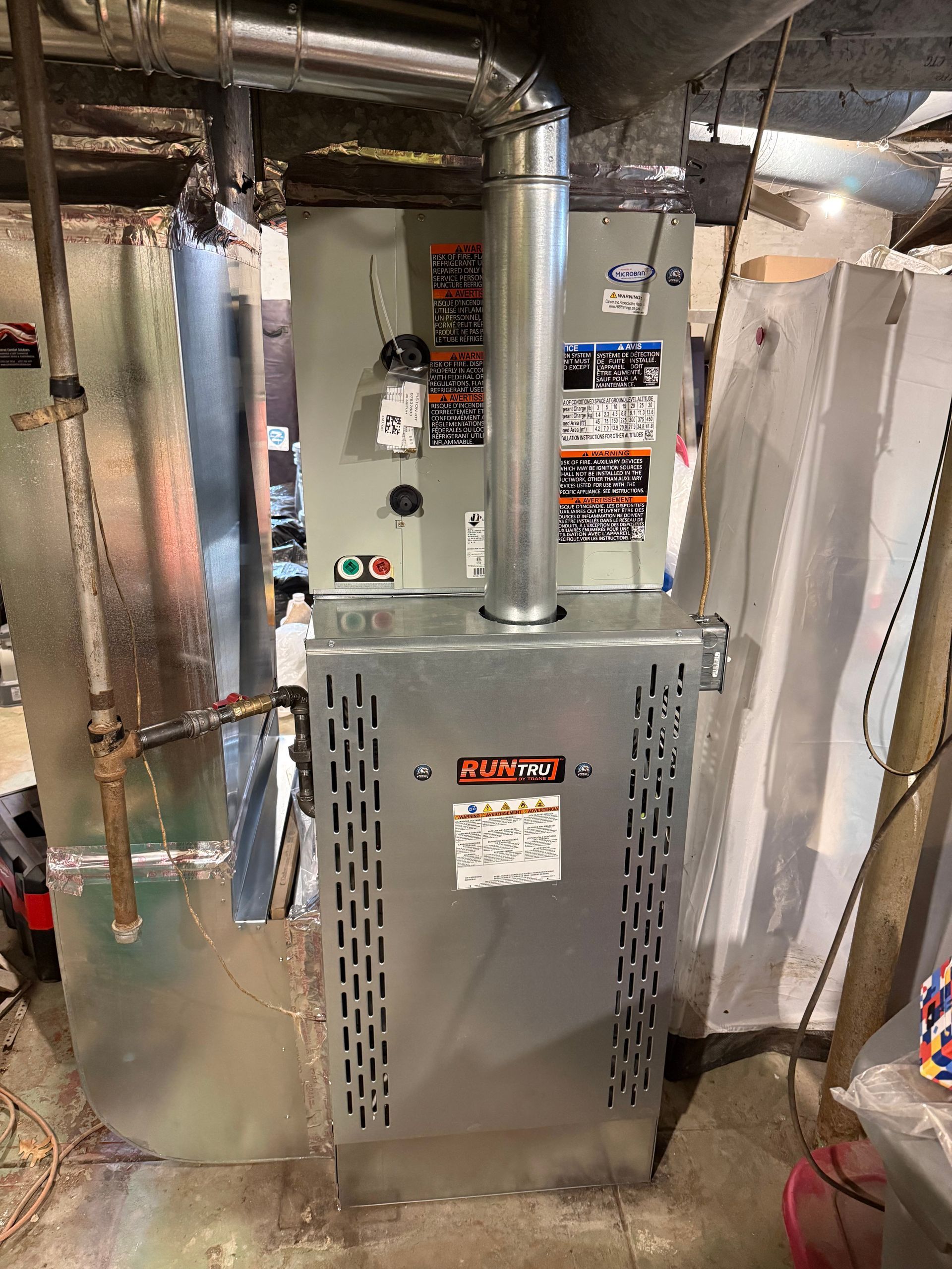 A gray HVAC furnace unit installed in a basement, connected to silver metal ductwork and a vertical exhaust pipe.