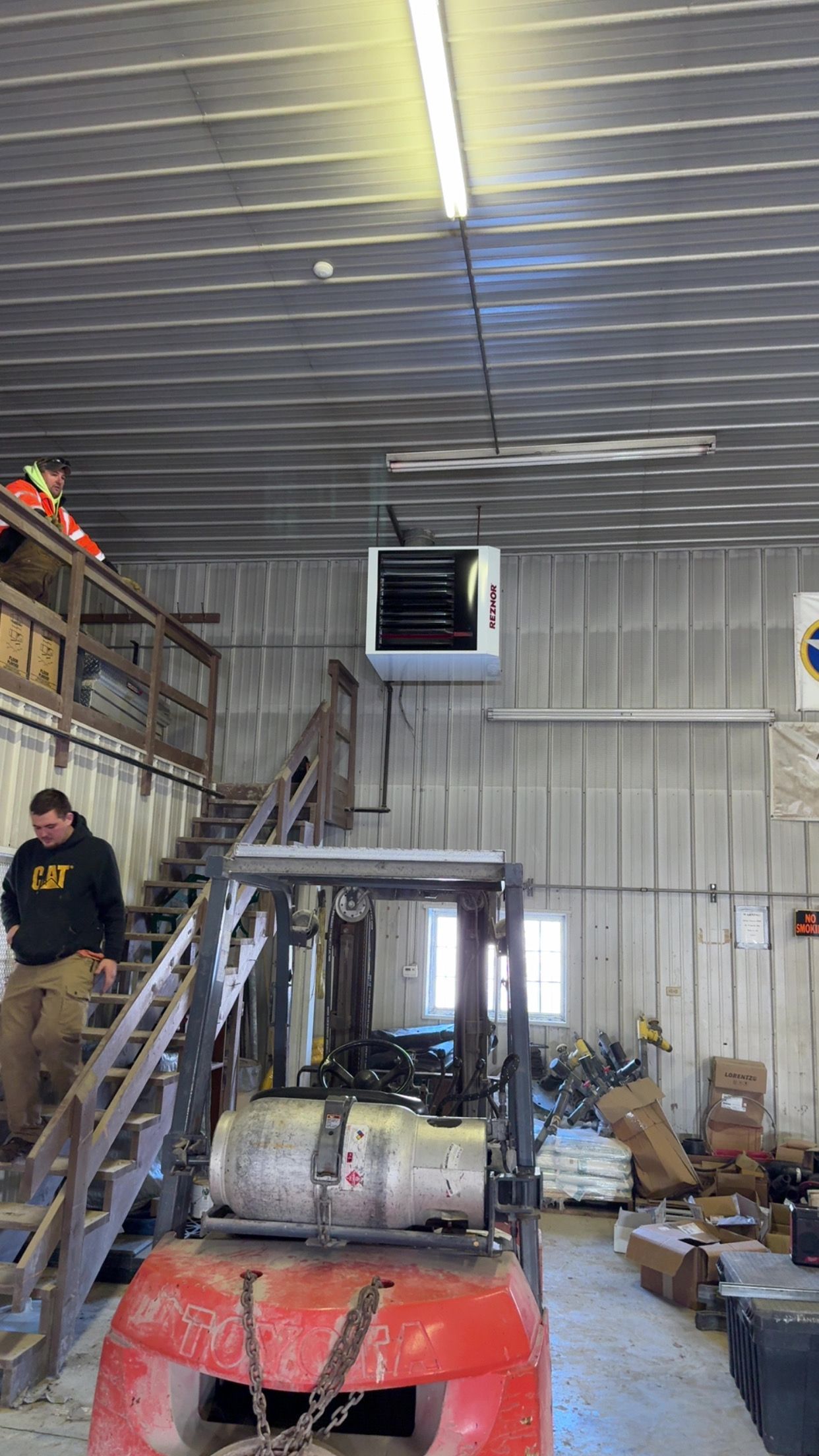 A person in a dark hoodie stands on stairs next to an orange forklift in a warehouse with a ceiling-mounted heater.