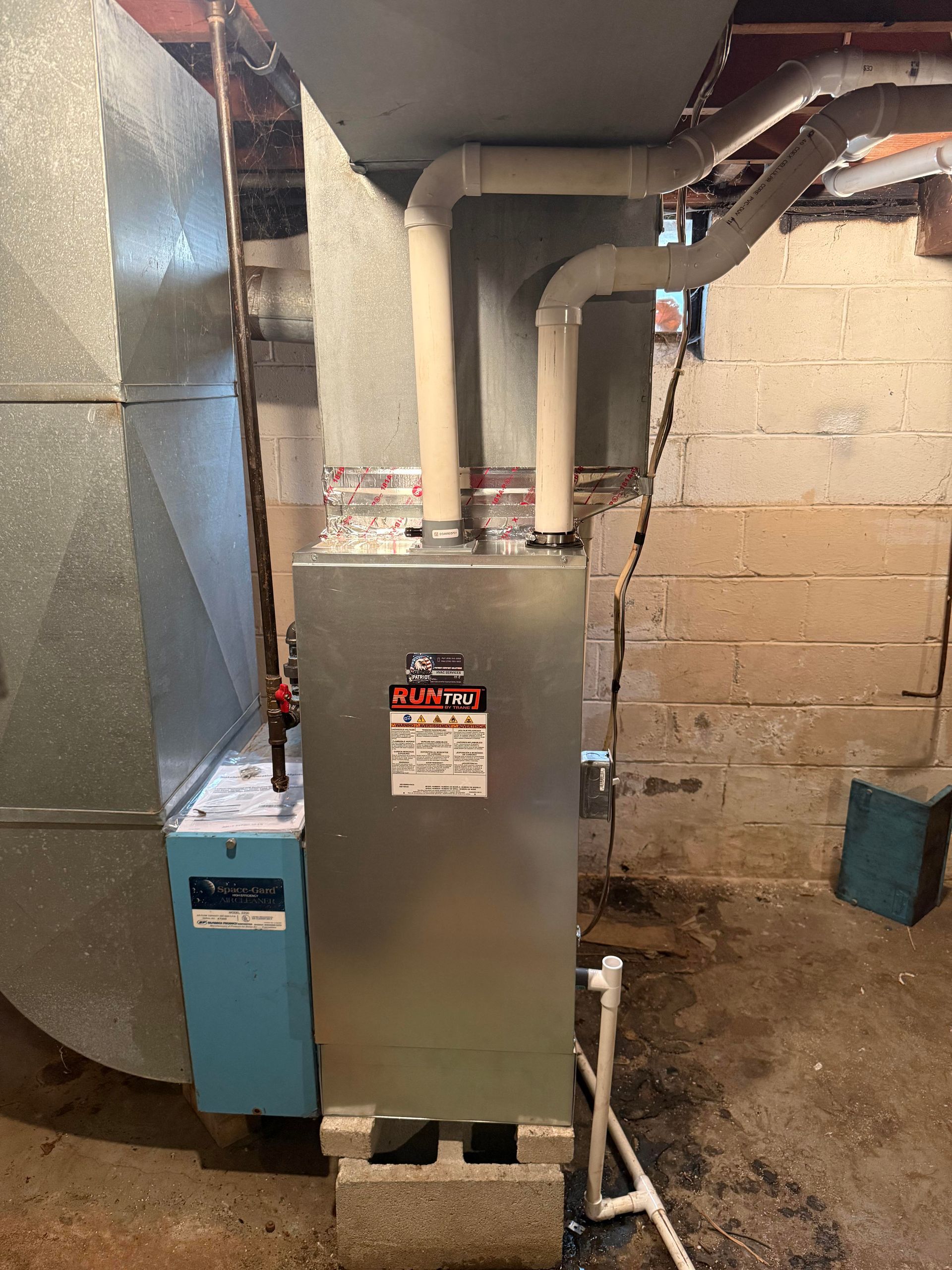 A furnace system with a silver metal unit, connected pipes, and a blue base, standing in a basement with concrete walls.