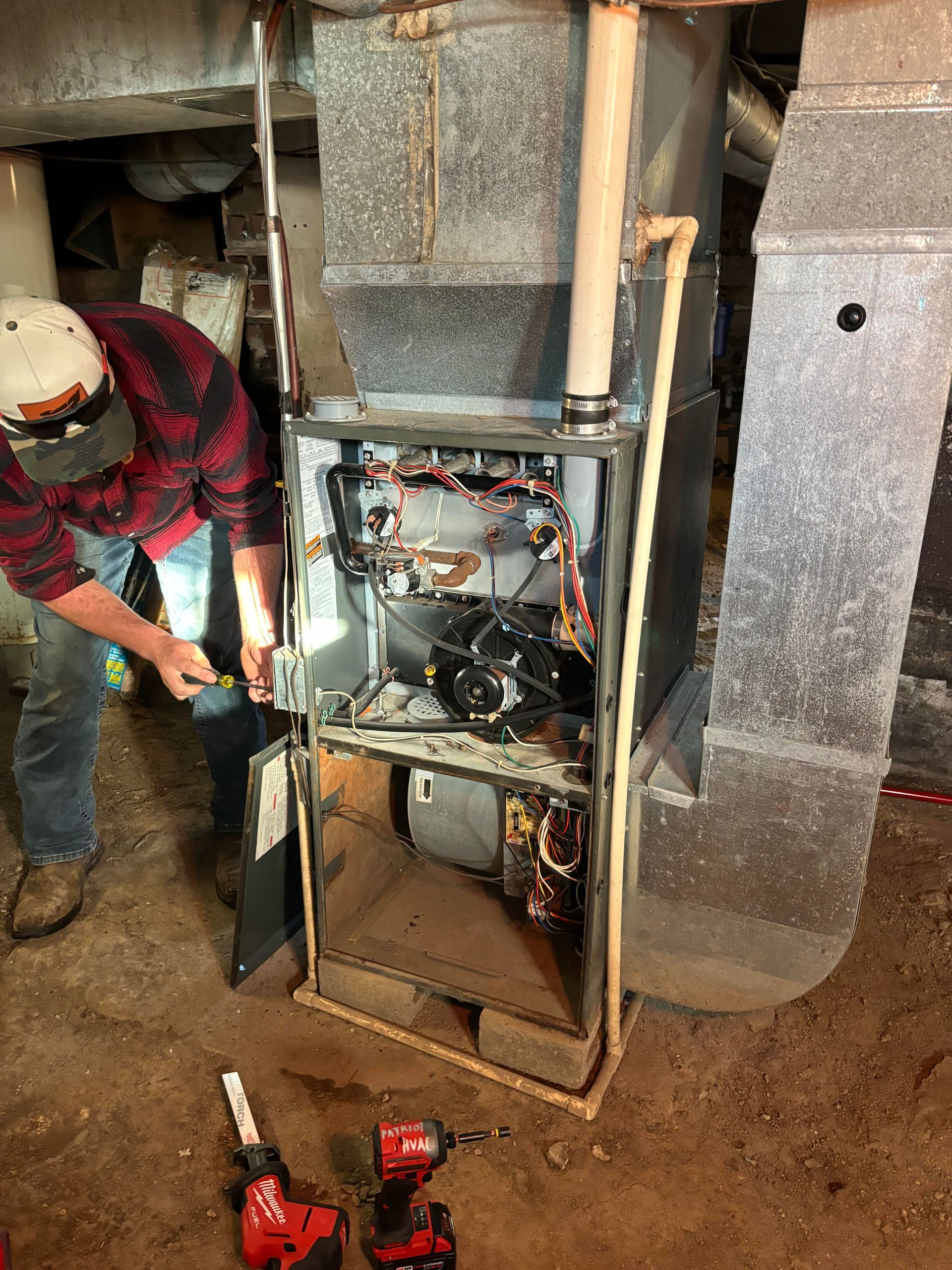 A technician in a red plaid shirt and baseball cap services an open furnace in a basement setting.