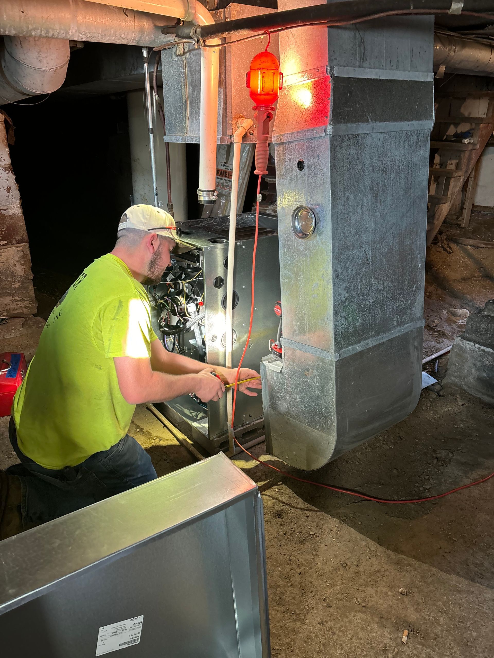 A technician wearing a neon yellow shirt works on a furnace in a basement with an orange work light hanging nearby.