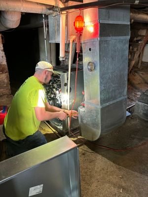 A technician in a lime green shirt repairs an HVAC furnace system in a dimly lit basement.