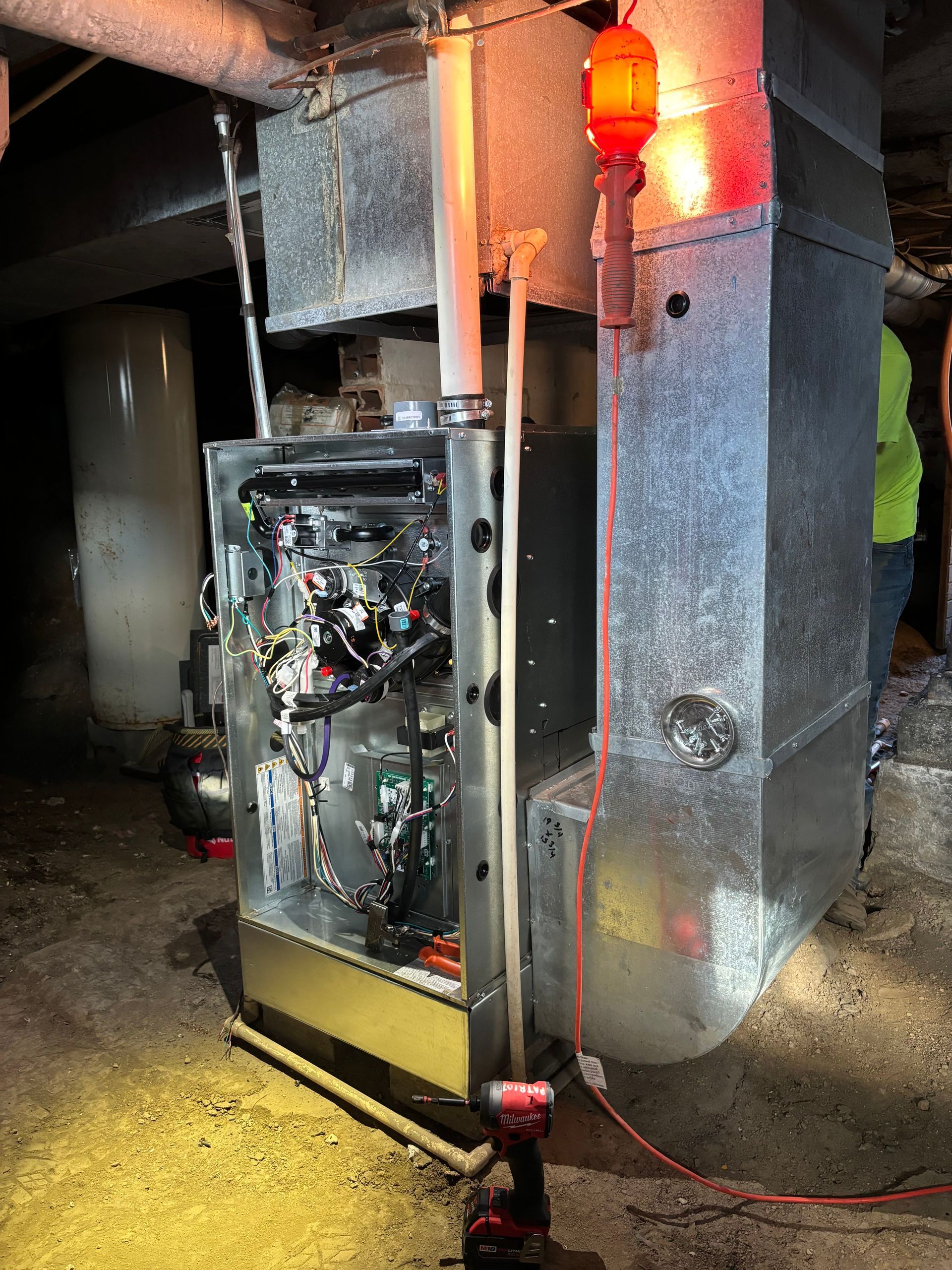 A gas furnace with an exposed front panel, standing in a basement, with an orange work light illuminating the ductwork.
