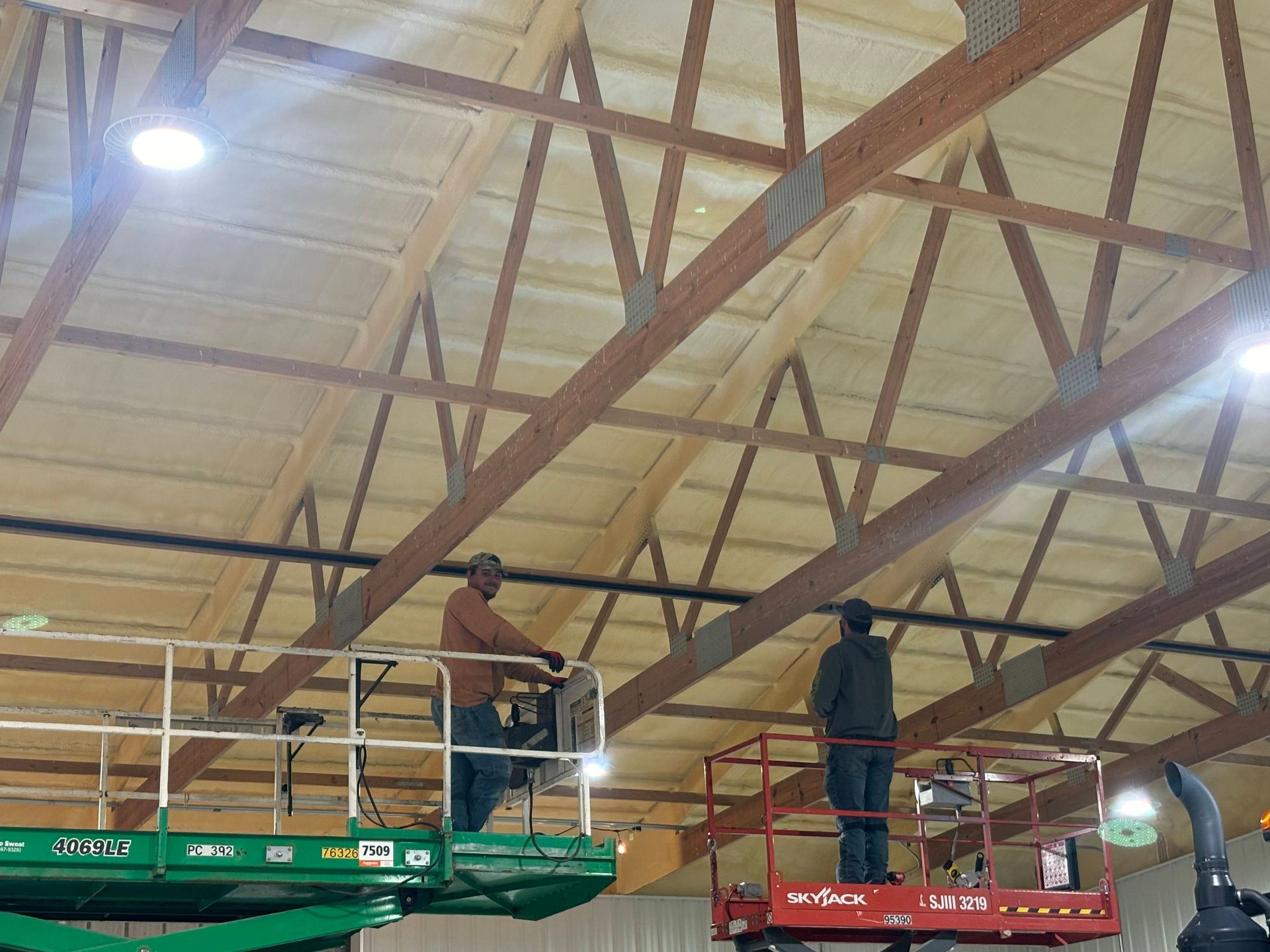 Two workers on raised scissor lifts perform maintenance on a large wooden truss ceiling inside a warehouse.