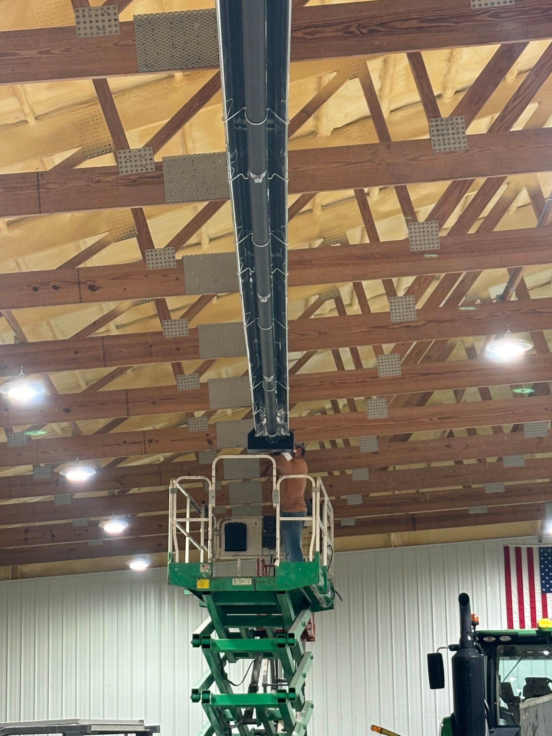 A person in a scissor lift works on a black, longitudinal structure mounted to the wooden trusses of a large building.