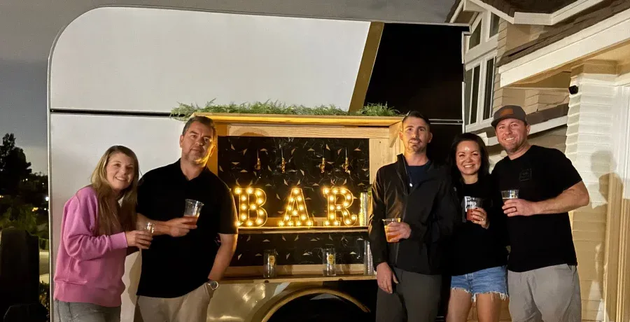 Six people pose with drinks near a bar trailer lit up with