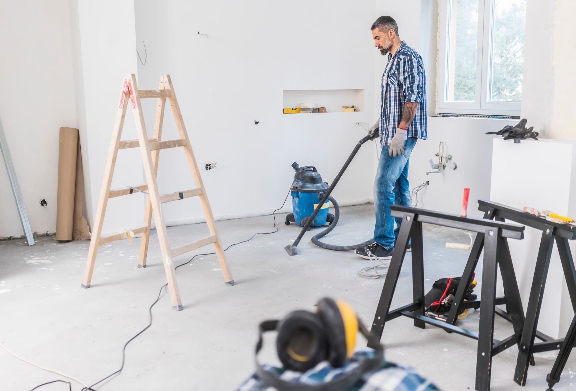 Man using a vacuum cleaner in a room under construction; a ladder and sawhorses are present.