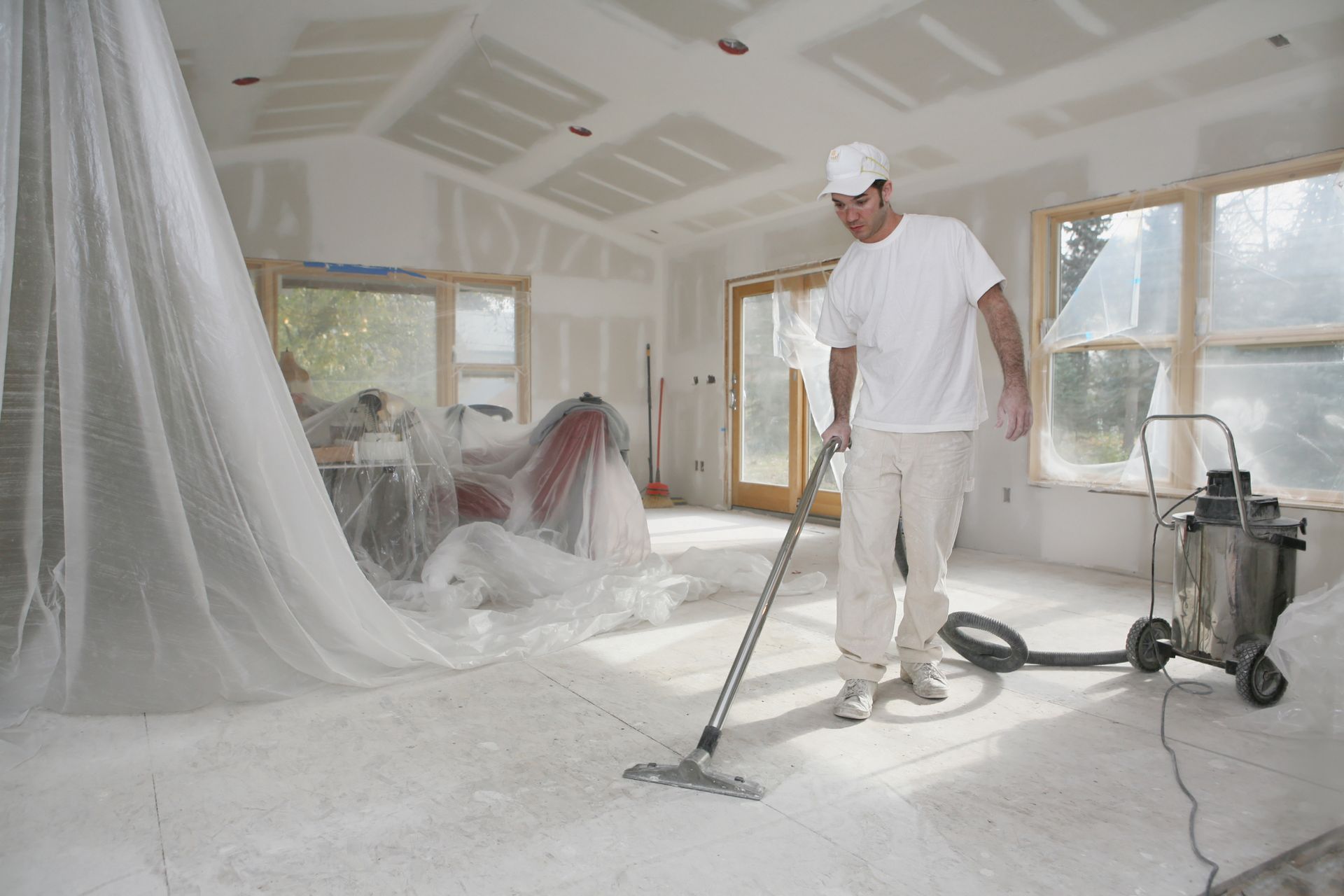 Man vacuums dust from floor of unfinished room with sheet-covered furniture and windows.