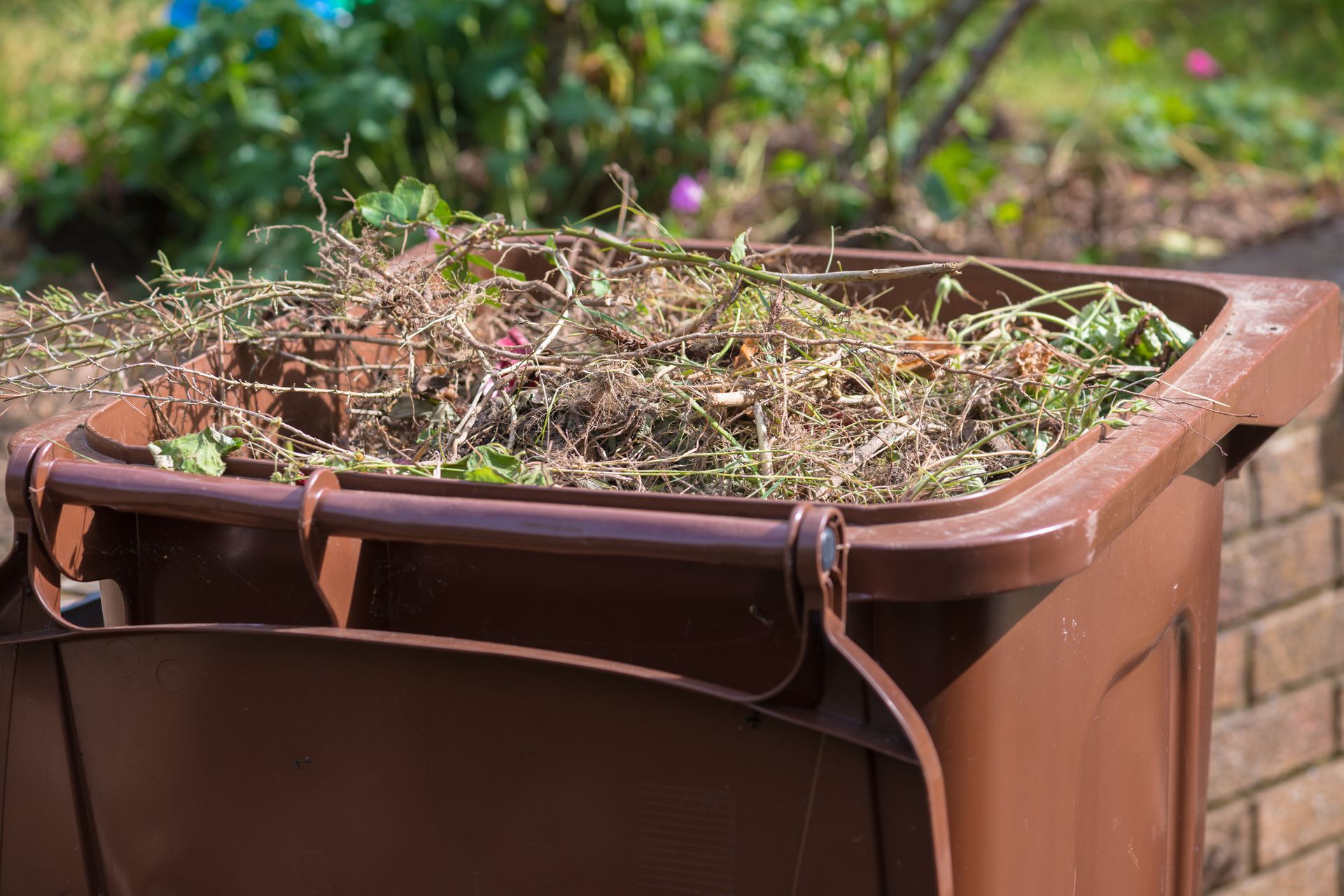 Brown trash bin filled with yard waste in a garden setting.
